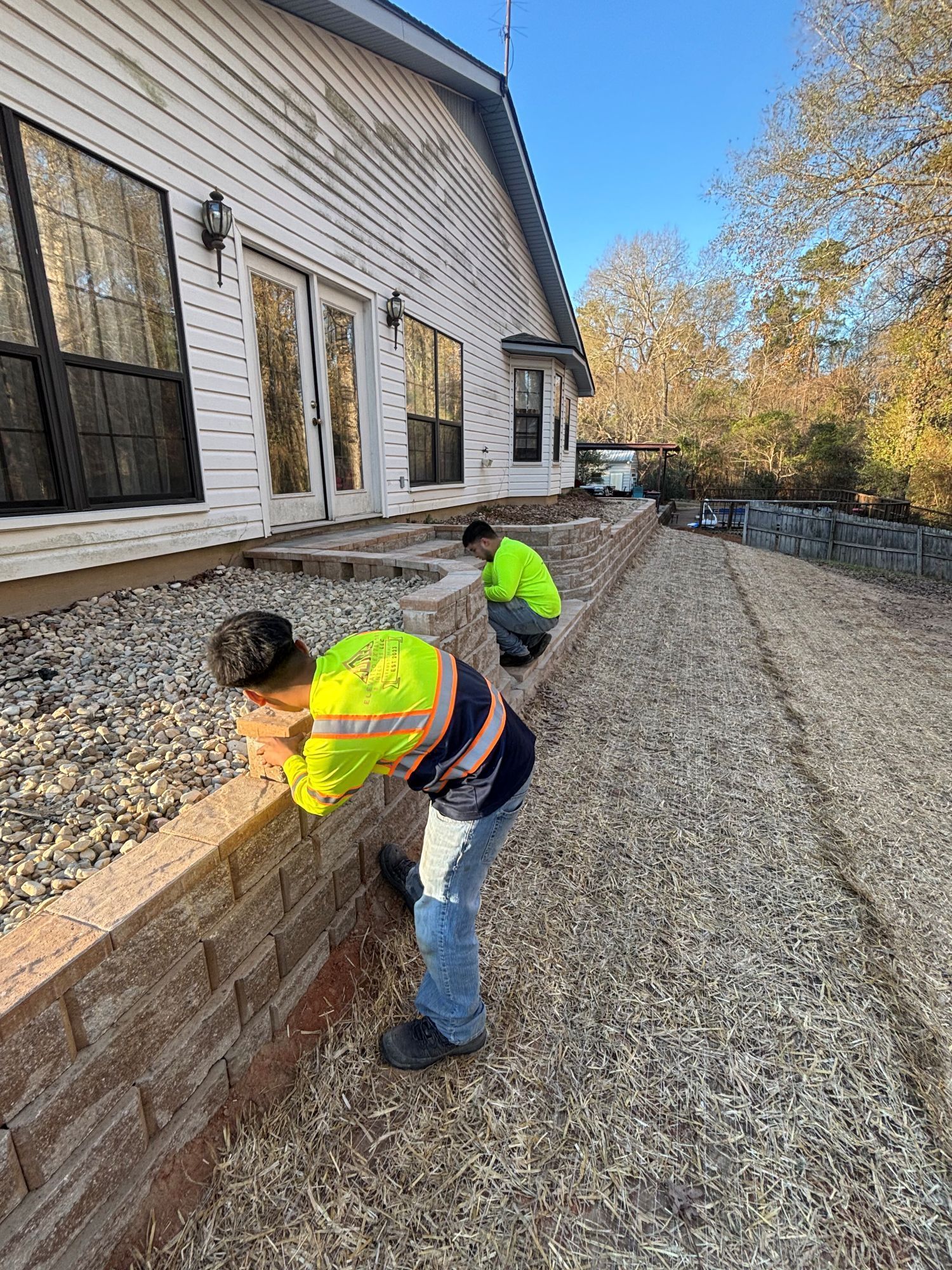 Two workers installing a retaining wall next to a house with gravel driveway. They wear safety vests.
