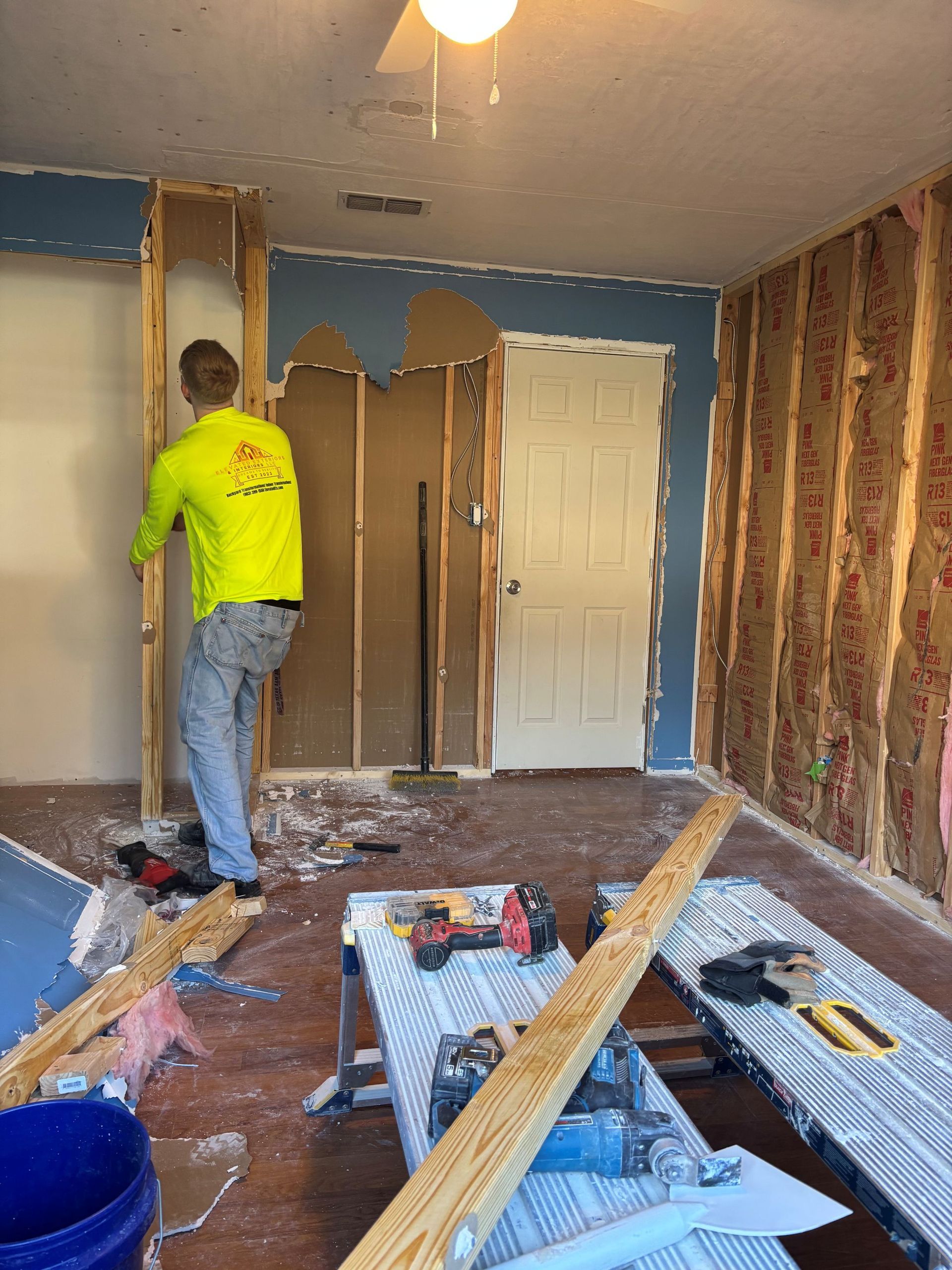 Man in yellow shirt demolishing wall in a room; tools and debris scattered around.