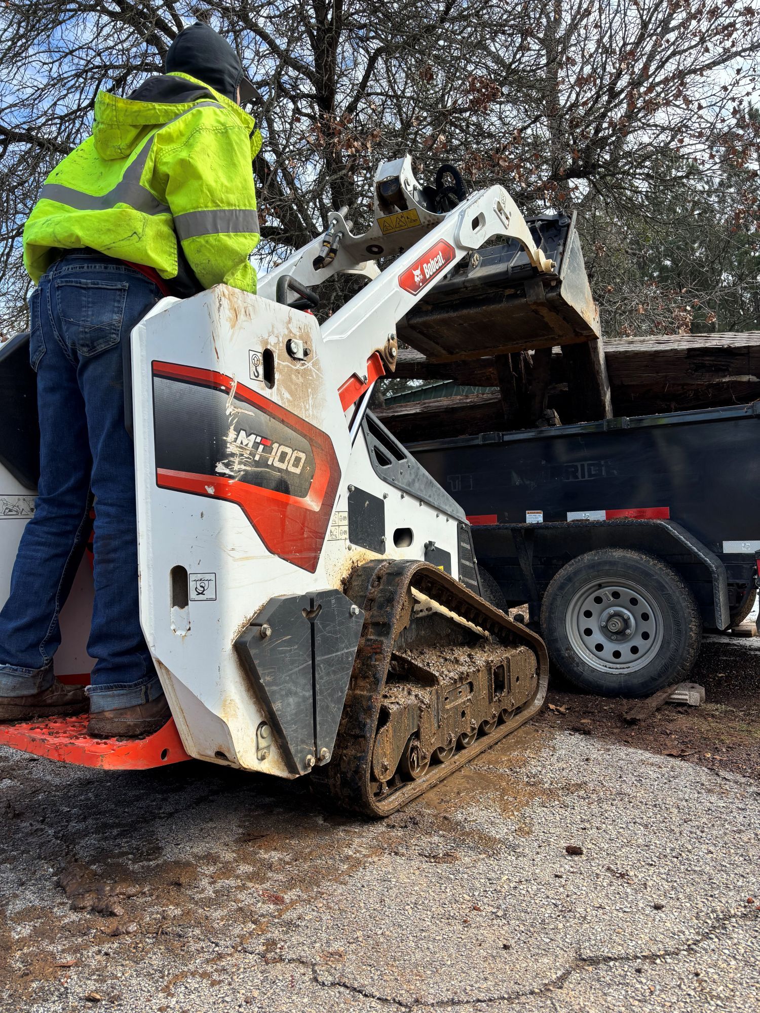 Person operating a Bobcat compact track loader, dumping into a trailer. They wear a neon yellow jacket.