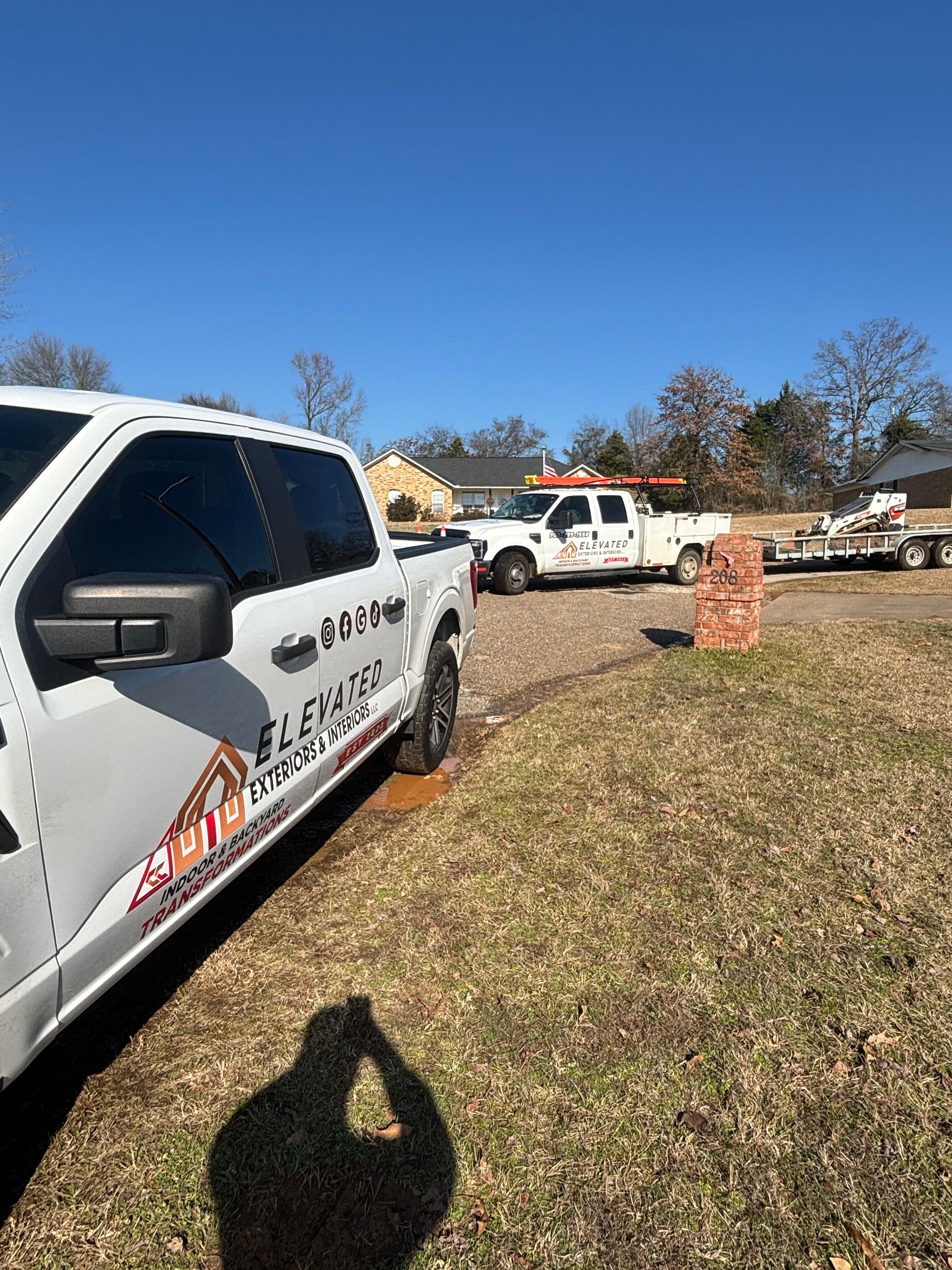 White work trucks on a grassy lot, sunny day. One truck close, other further back with a trailer.