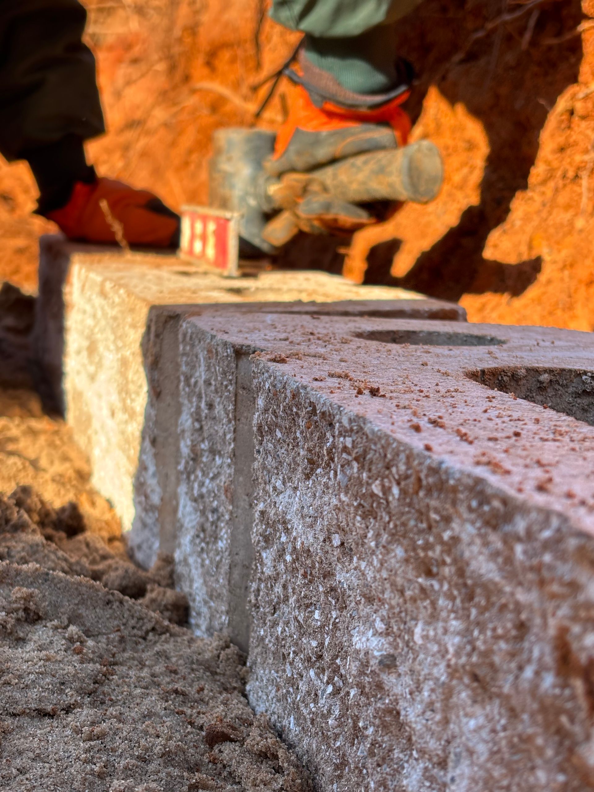 A person levels a brick with a mallet. Construction site with reddish-brown earth and tan bricks.