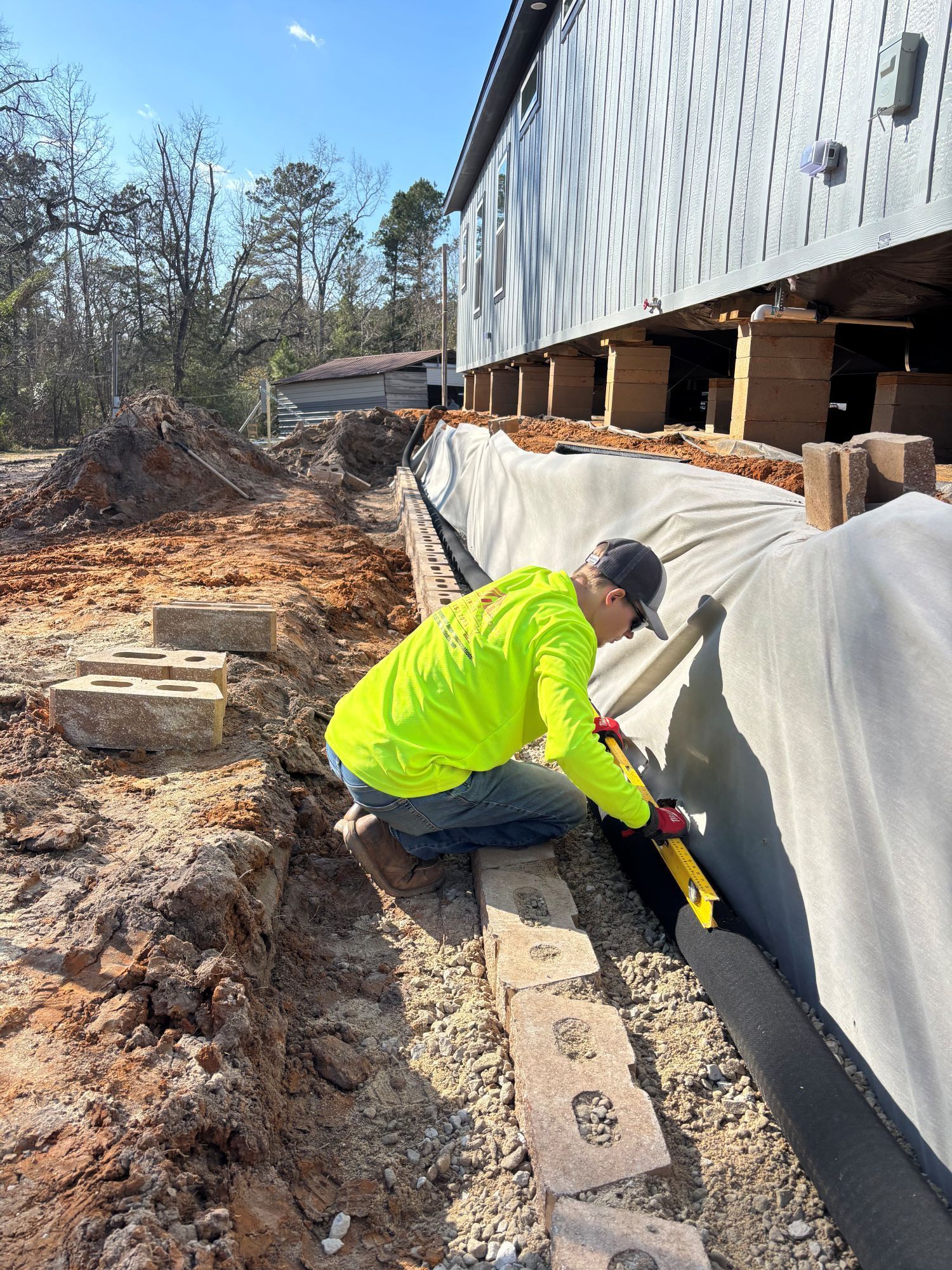 Man in neon jacket cuts fabric near a building's foundation, likely for a landscaping project.
