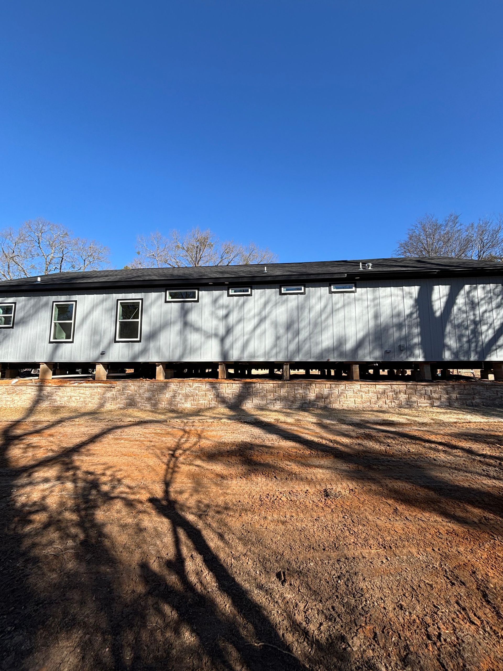 Long, gray building with small windows, raised on supports, under a blue sky, with tree shadows in the foreground.