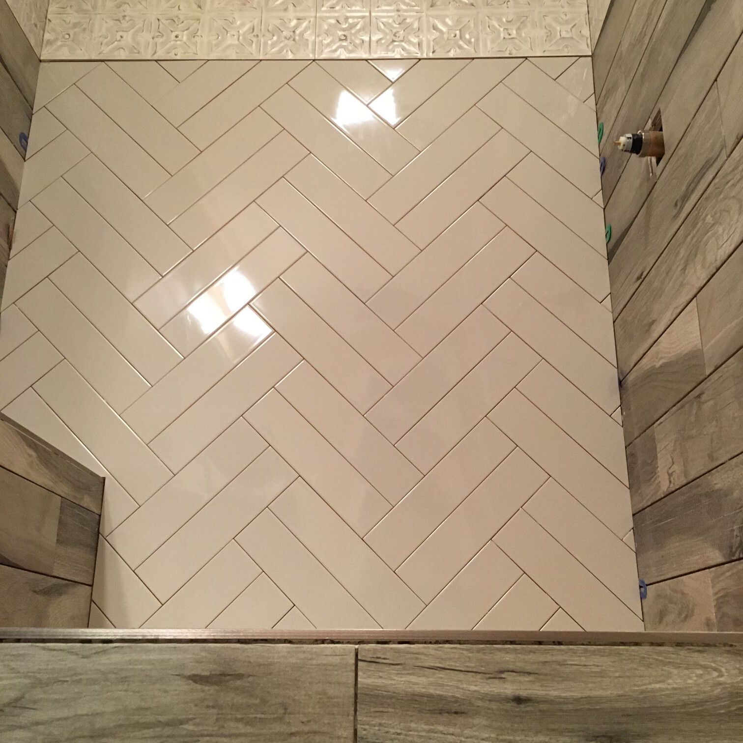 Herringbone pattern tile floor in a shower. White rectangular tiles. Wood-look tile on surrounding walls.