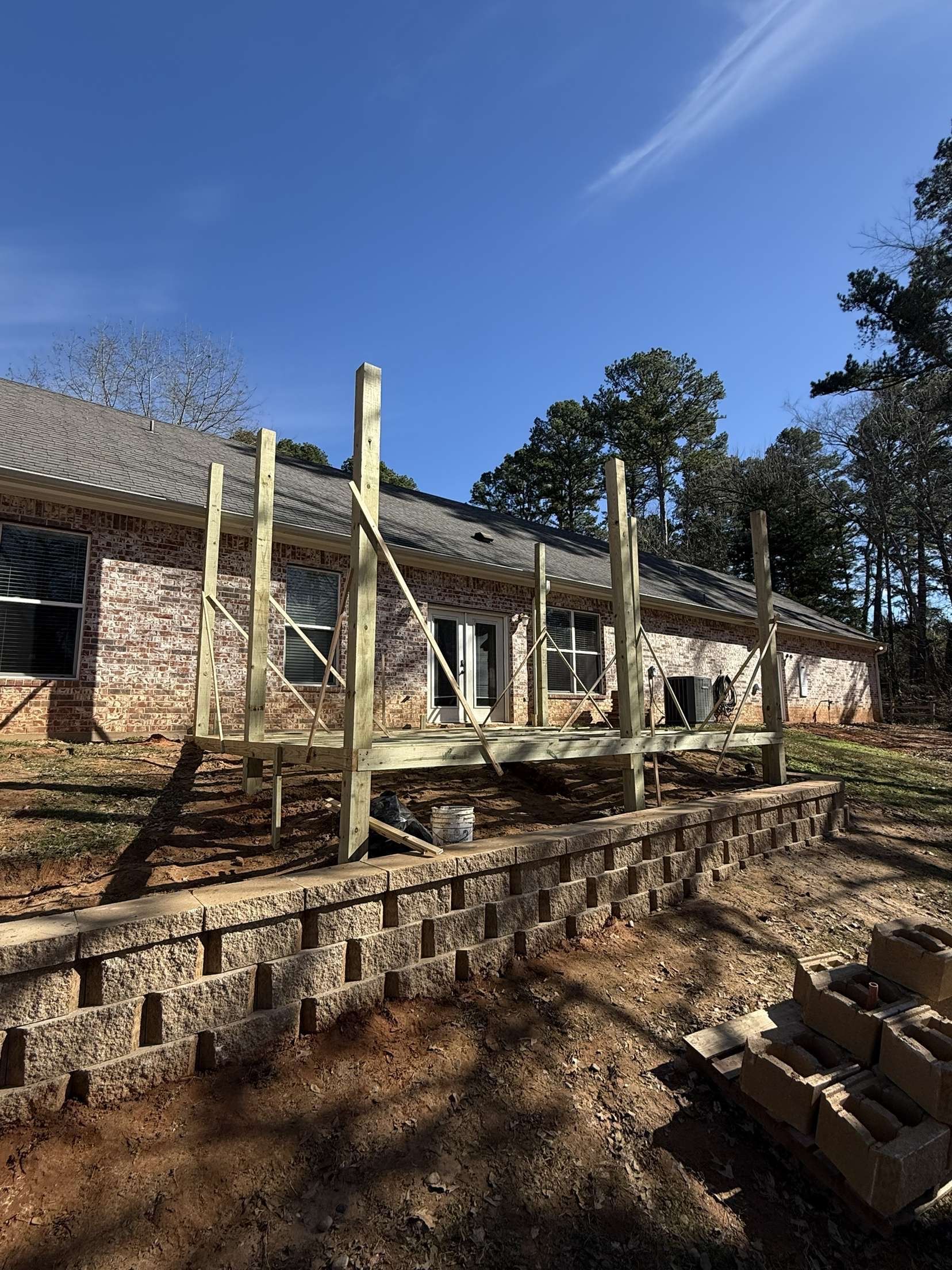 Wooden deck under construction next to a brick house and a retaining wall, on a sunny day.