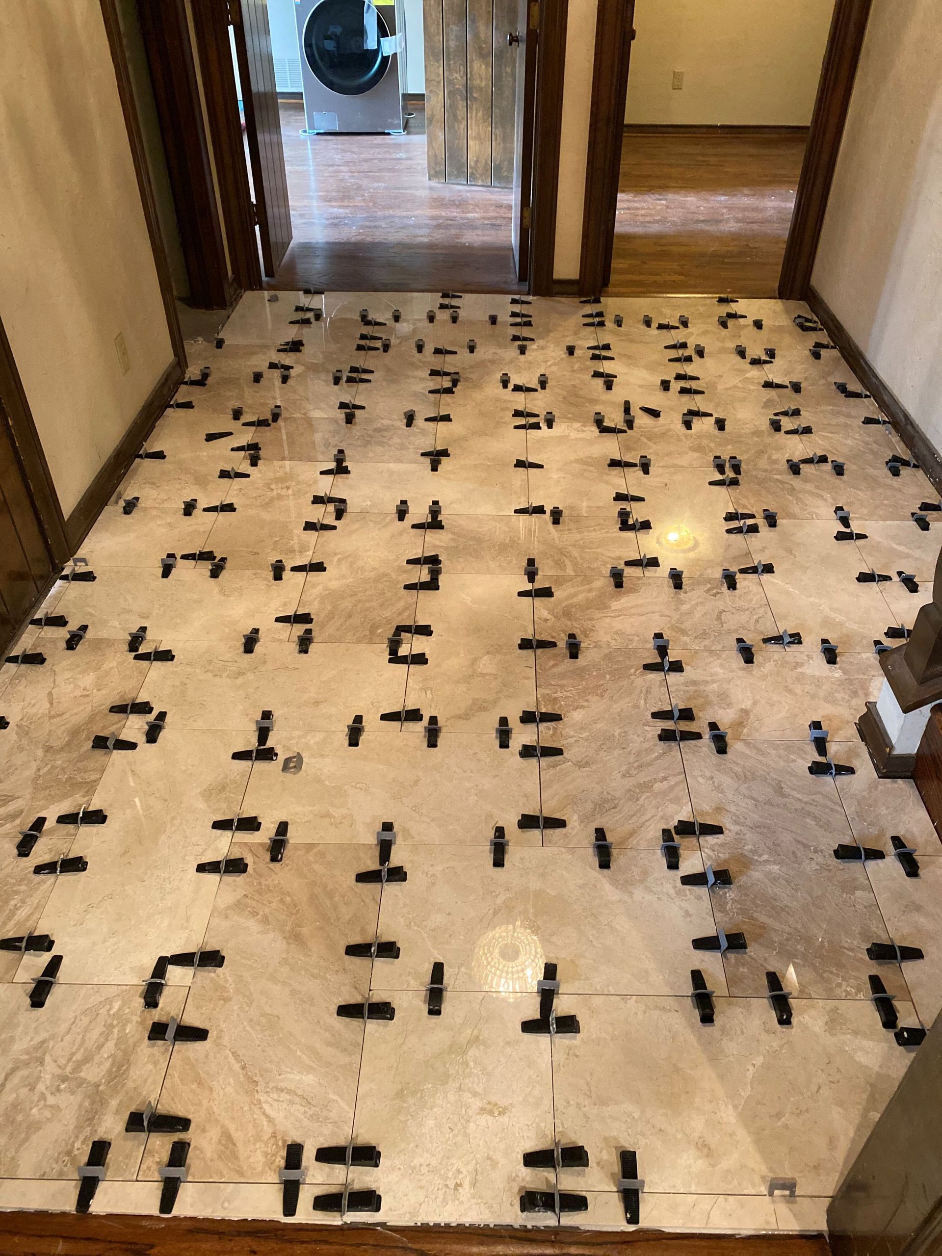 Tiled floor installation with leveler clips. Beige tiles in a hallway, door in background.
