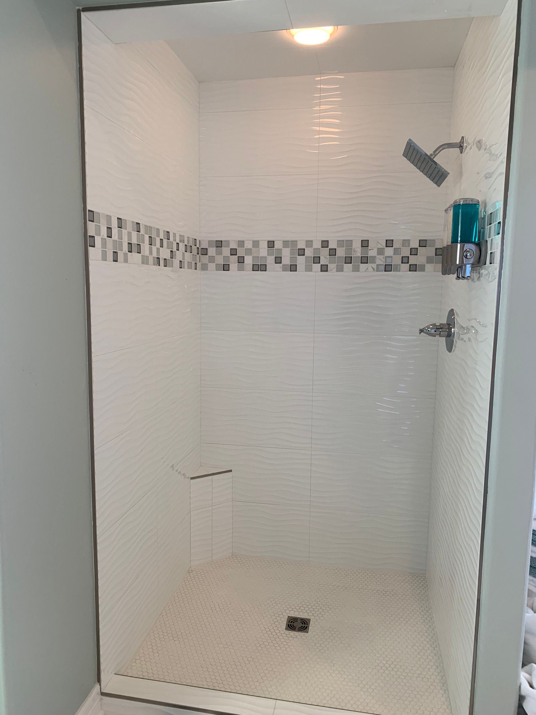 White tiled shower with mosaic tile stripe, showerhead, and soap dispenser.