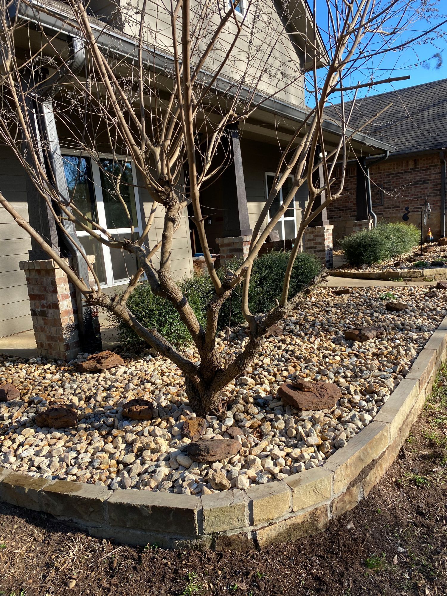 Bare tree in a rock bed in front of a house with a porch and brick accents. Sunny day.