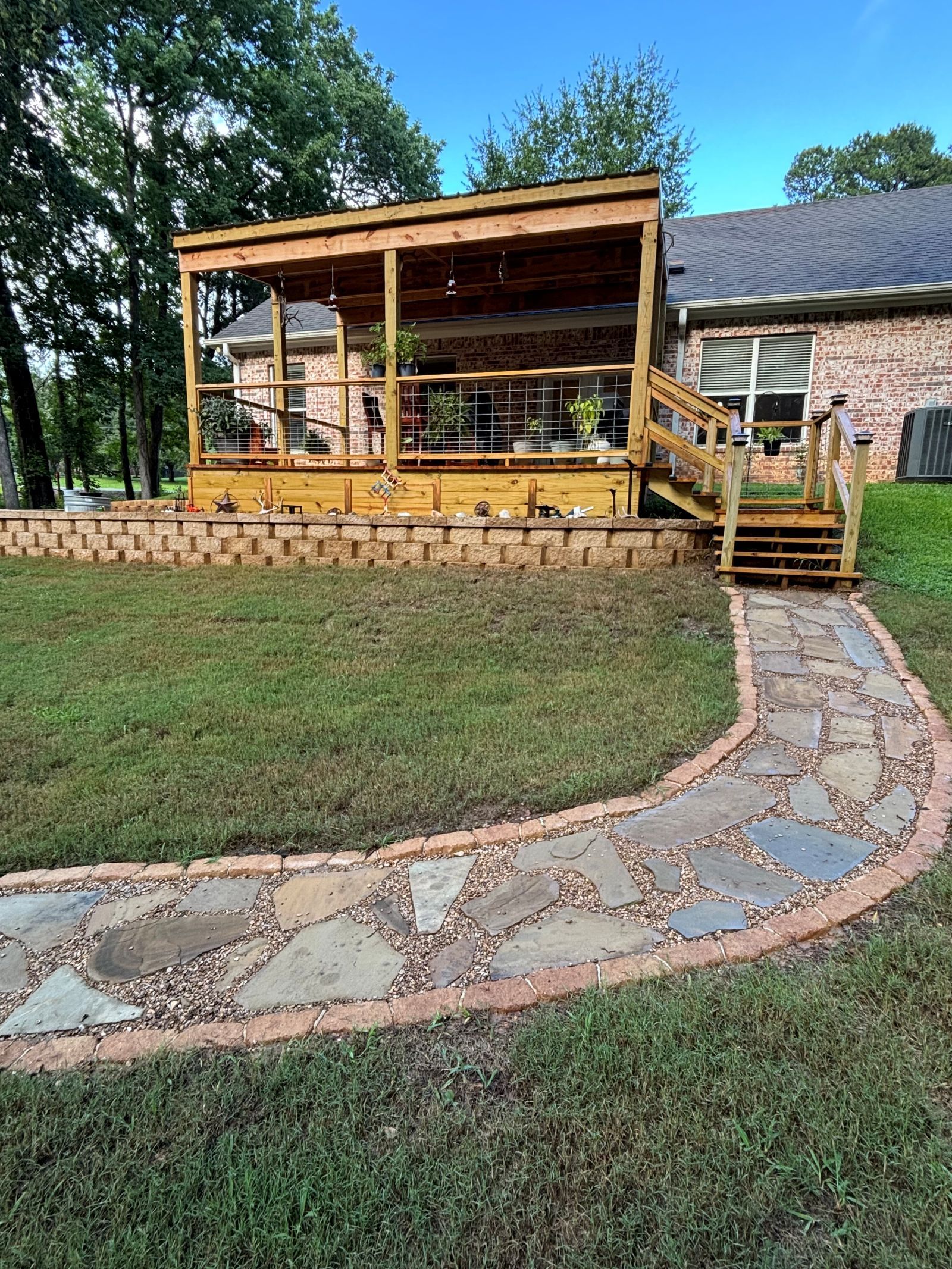Stone path leads to a wooden deck with a pergola, brick house in the background. Green grass and trees surround.