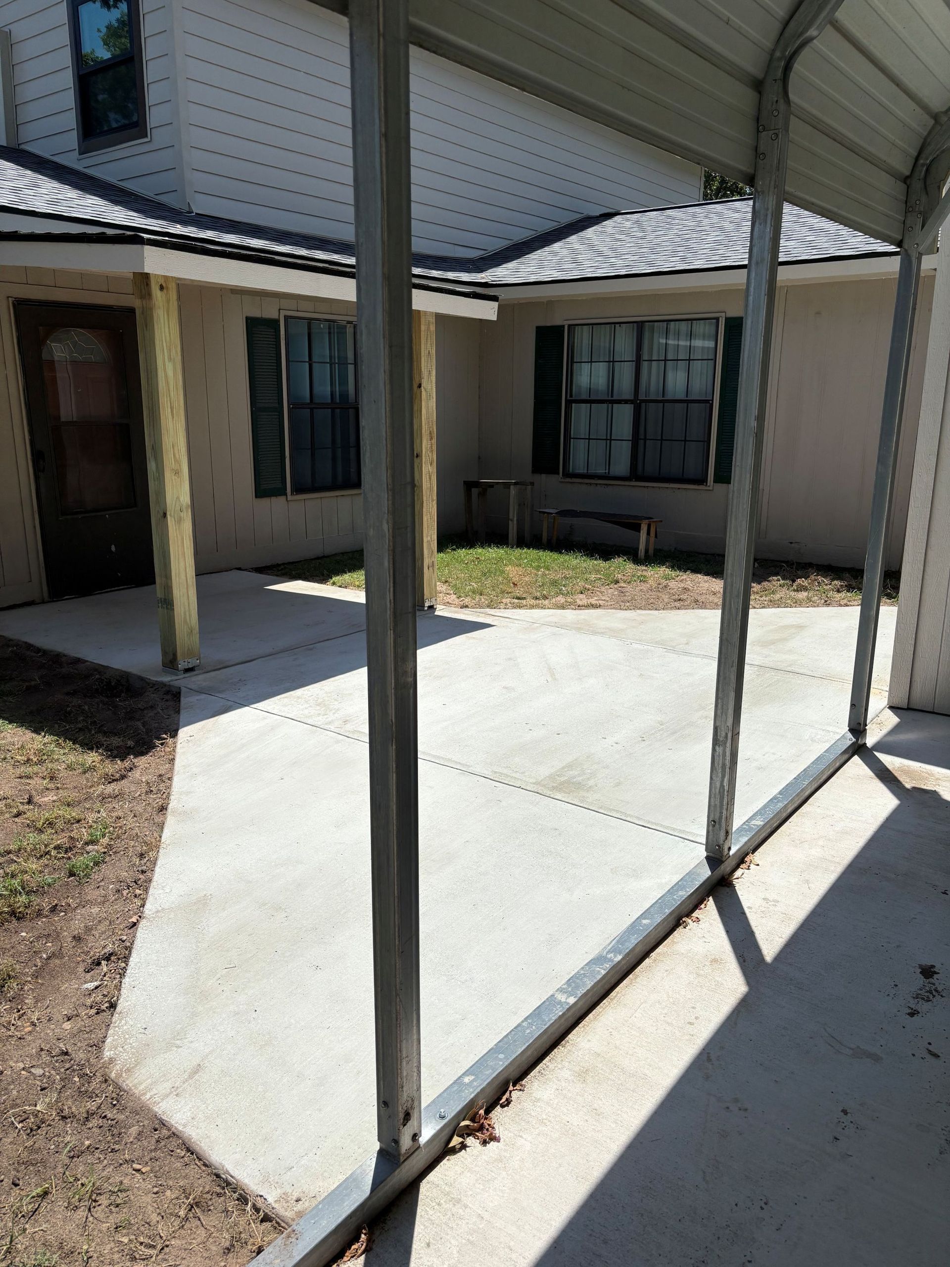 Concrete patio with metal awning attached to a beige house, with a wooden door and green shutters.