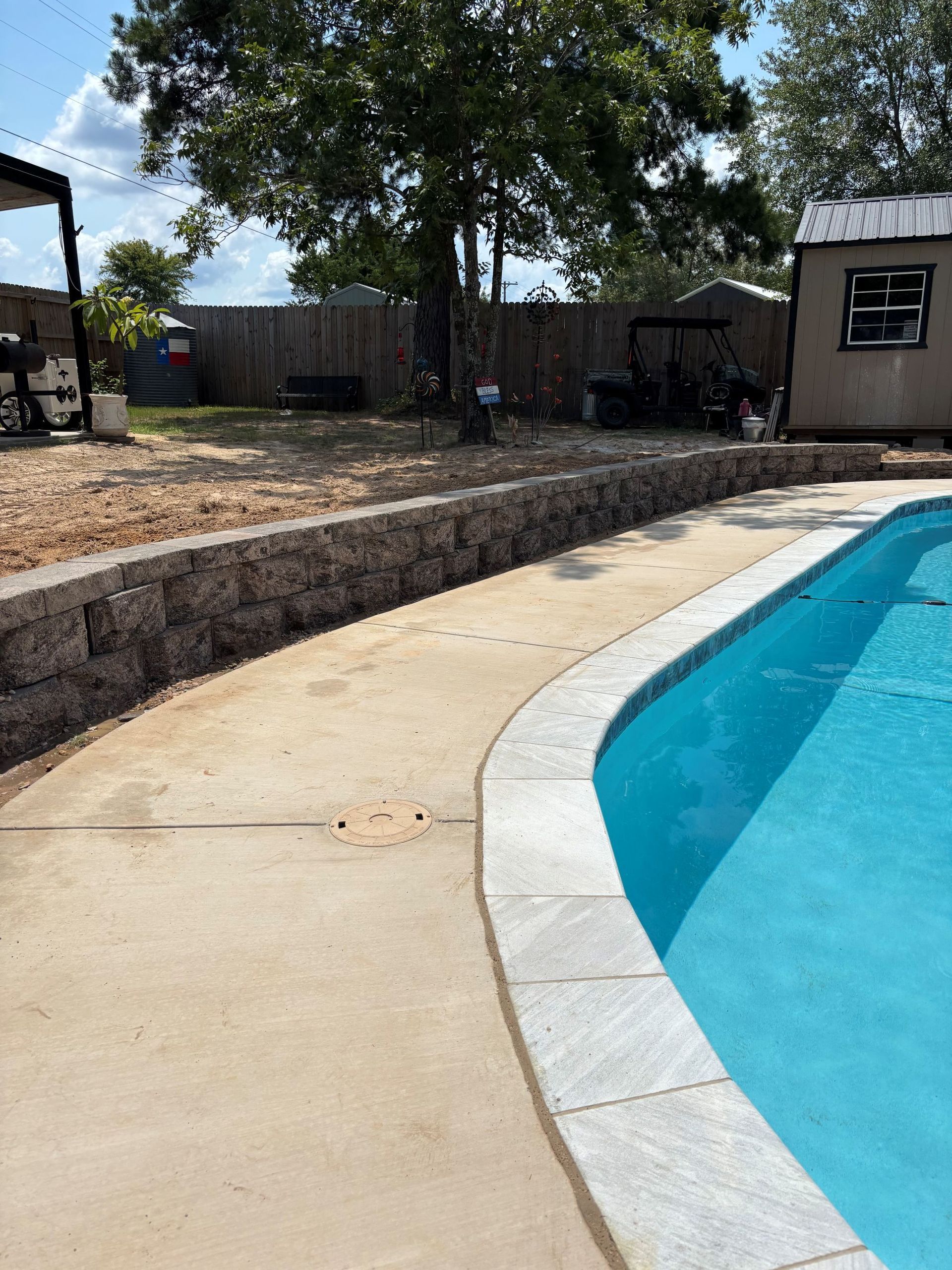 Concrete pool deck next to a pool. A retaining wall of stacked stones borders a dirt area.