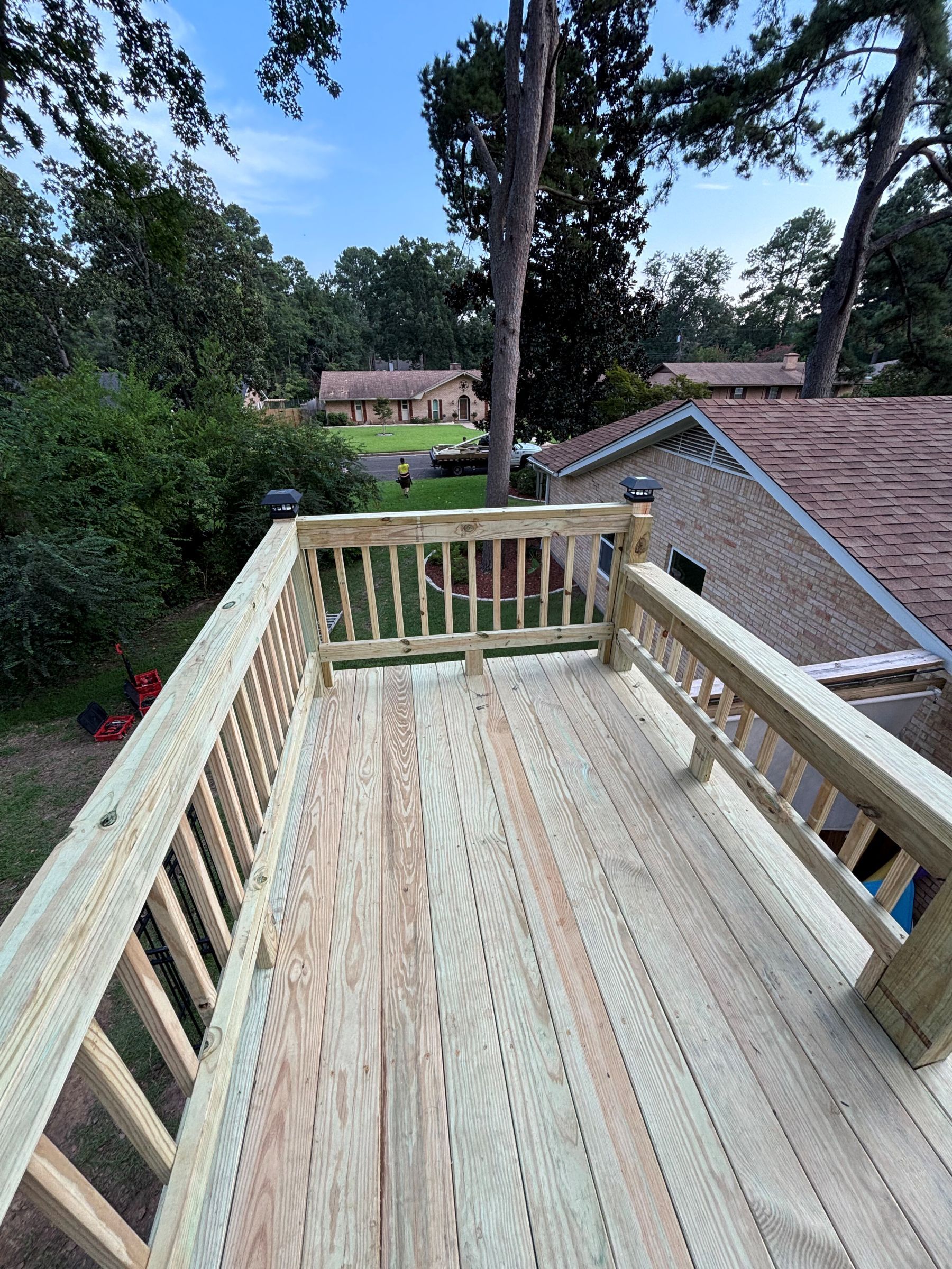Wooden deck with railing, overlooking a yard with trees and a brick house.