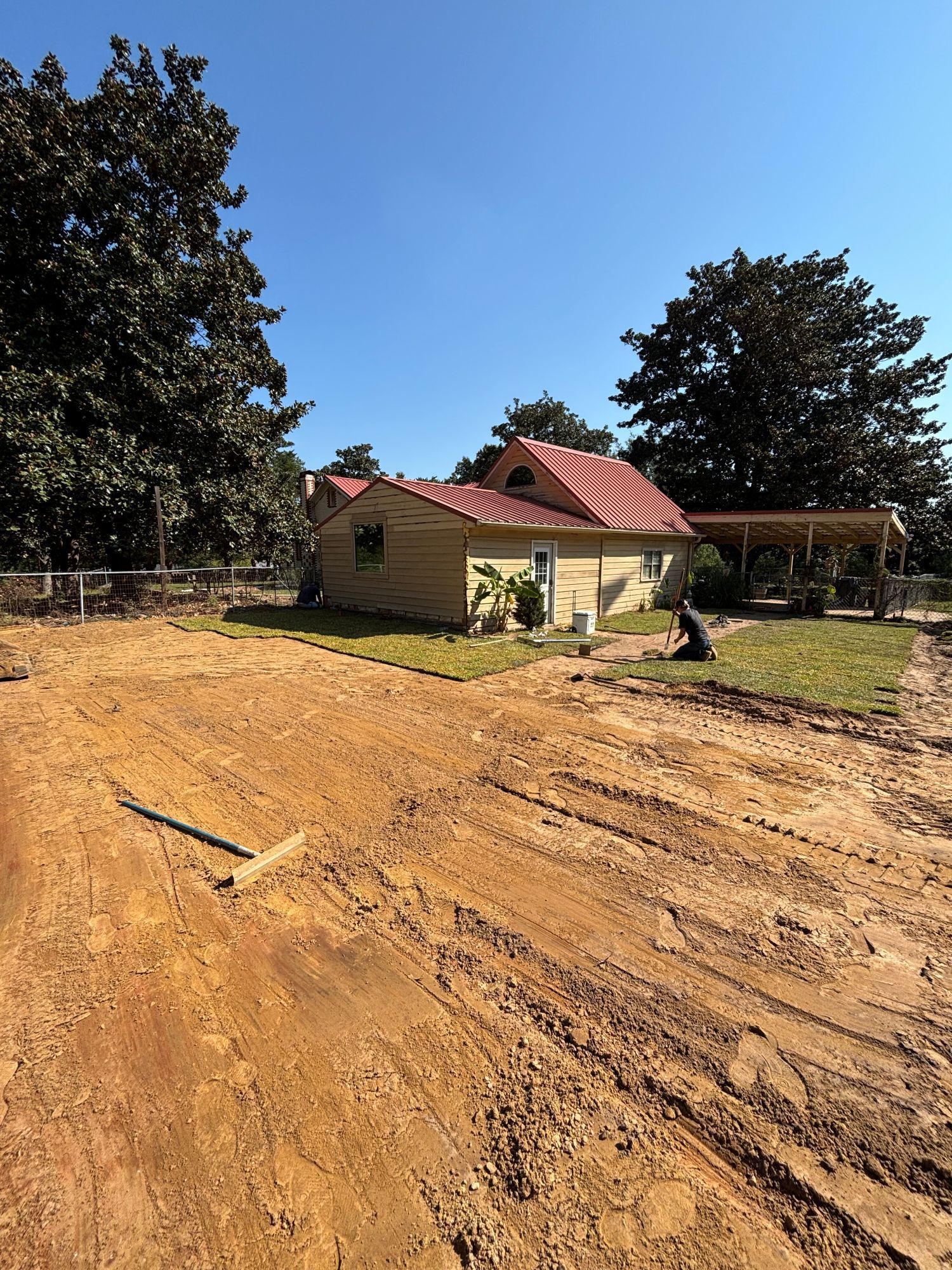 Dirt lot in front of a tan building with a red roof, a person sitting near the building, and trees in the background.