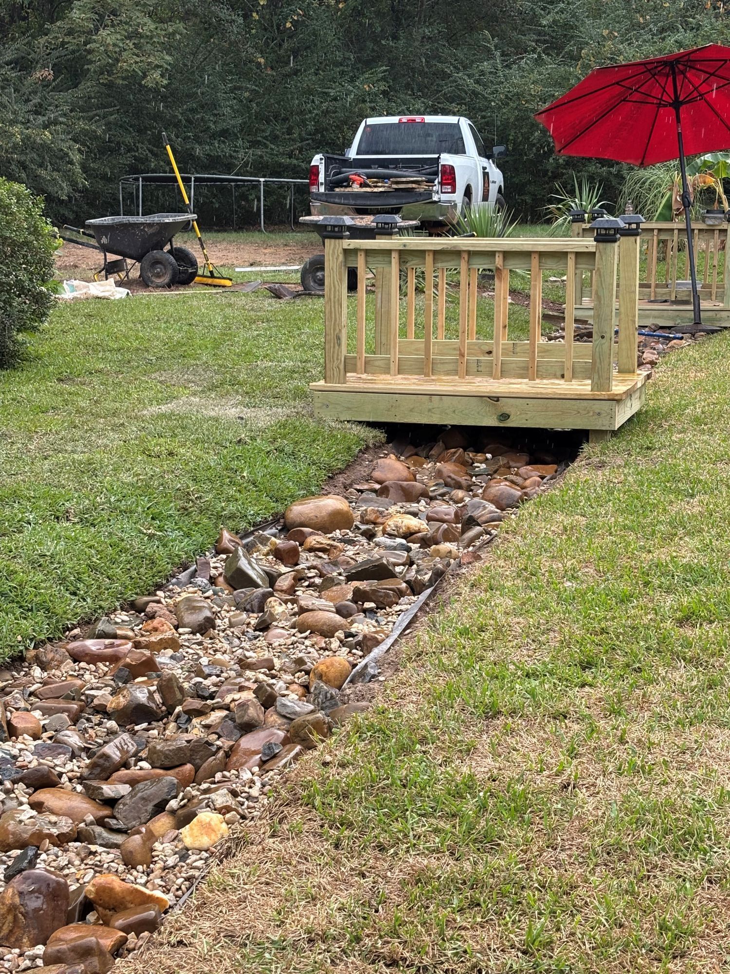 A wooden bridge over a rock-filled creek in a grassy yard, with a truck and umbrella visible.