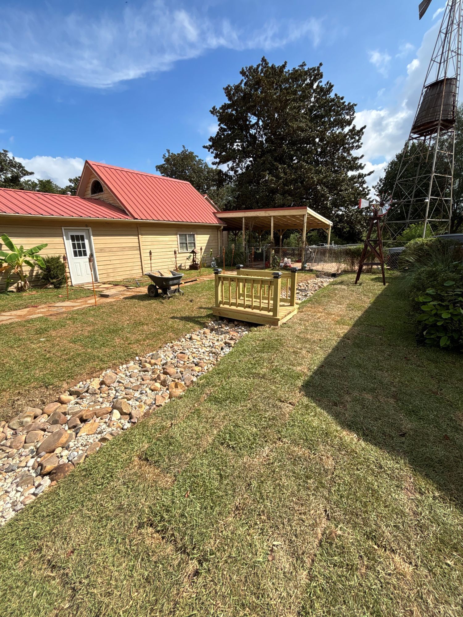 Yellow house with a red roof, small wooden bridge over a rock-lined drainage ditch, sunny day.