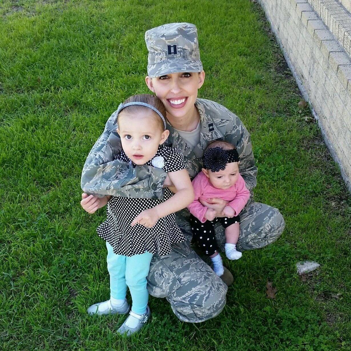 A woman in a military uniform is kneeling down holding two babies