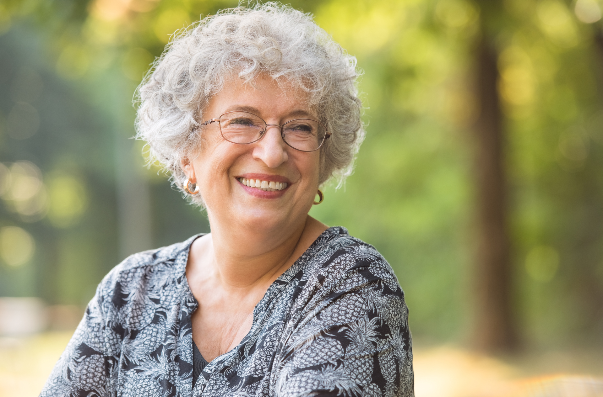 An elderly woman with glasses is smiling in a park.