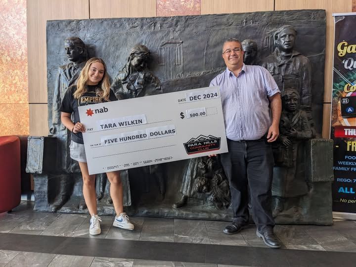 A man and a woman are holding a large check in front of a statue at Para Hills Community Club