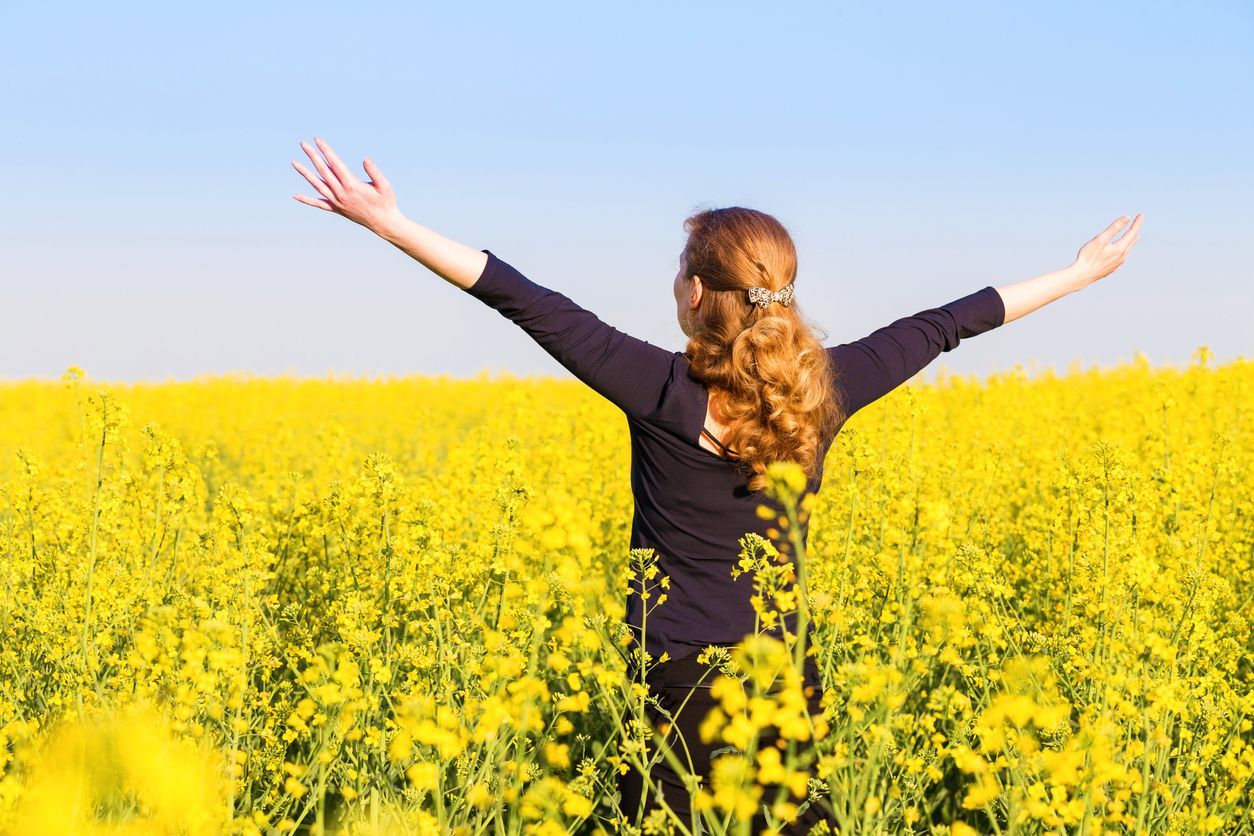 Woman with arms outstretched in a field of yellow flowers under a blue sky.