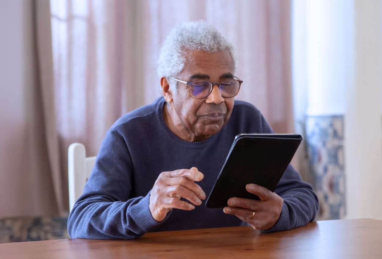 Older Black man with glasses using a tablet at a wooden table. Indoors, looking focused.