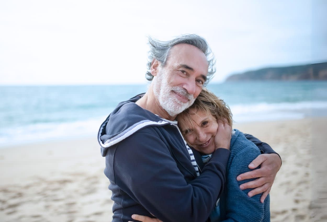 Older couple hugging on a beach, smiling at the camera. Blue sea in the background.