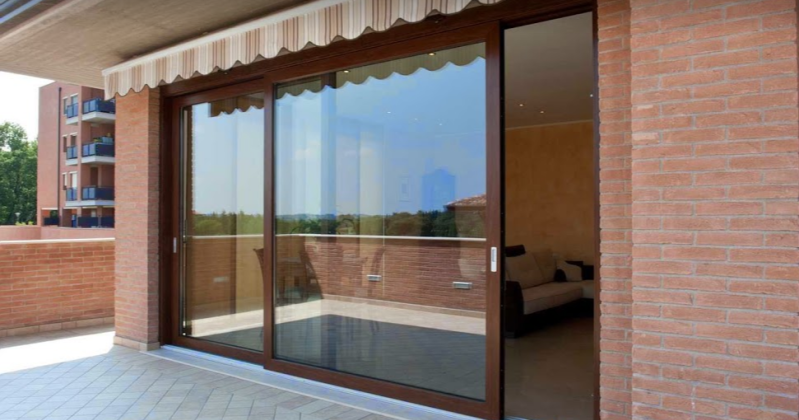 Porte scorrevoli in vetro con telai marroni su un balcone con mattoni rossi e tenda da sole.