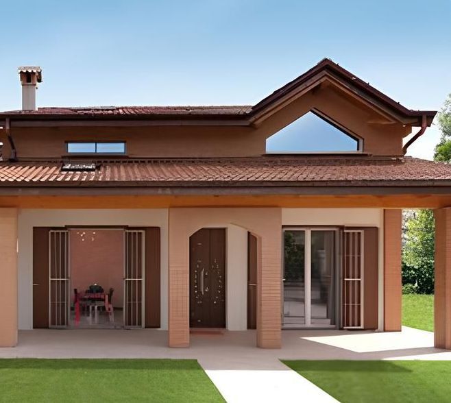 Casa a due piani marrone e beige con tetto a due falde, veranda e persiane, su un prato.