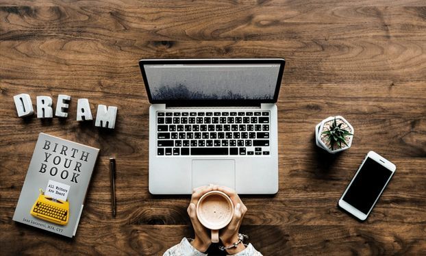 A woman’s hands holding a cup of coffee in front of her opened laptop in the center, DREAM in white block letters on the top left, the Birth Your Book workbook/teaching memoir on the bottom left with a bright yellow typewriter reading “All Writers Are Heard”, a small succulent and cell phone on the right. They are all sitting atop a dark brown wooden desk