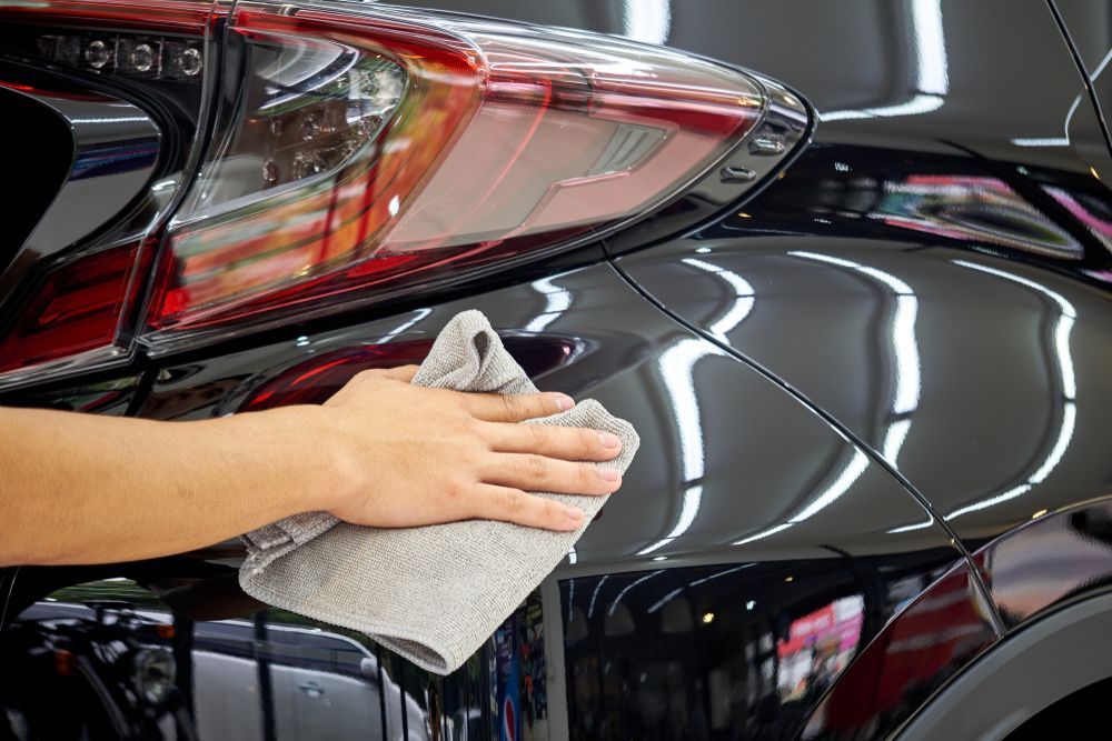 Hand wiping the glossy black car exterior with a gray microfiber cloth near a red taillight.