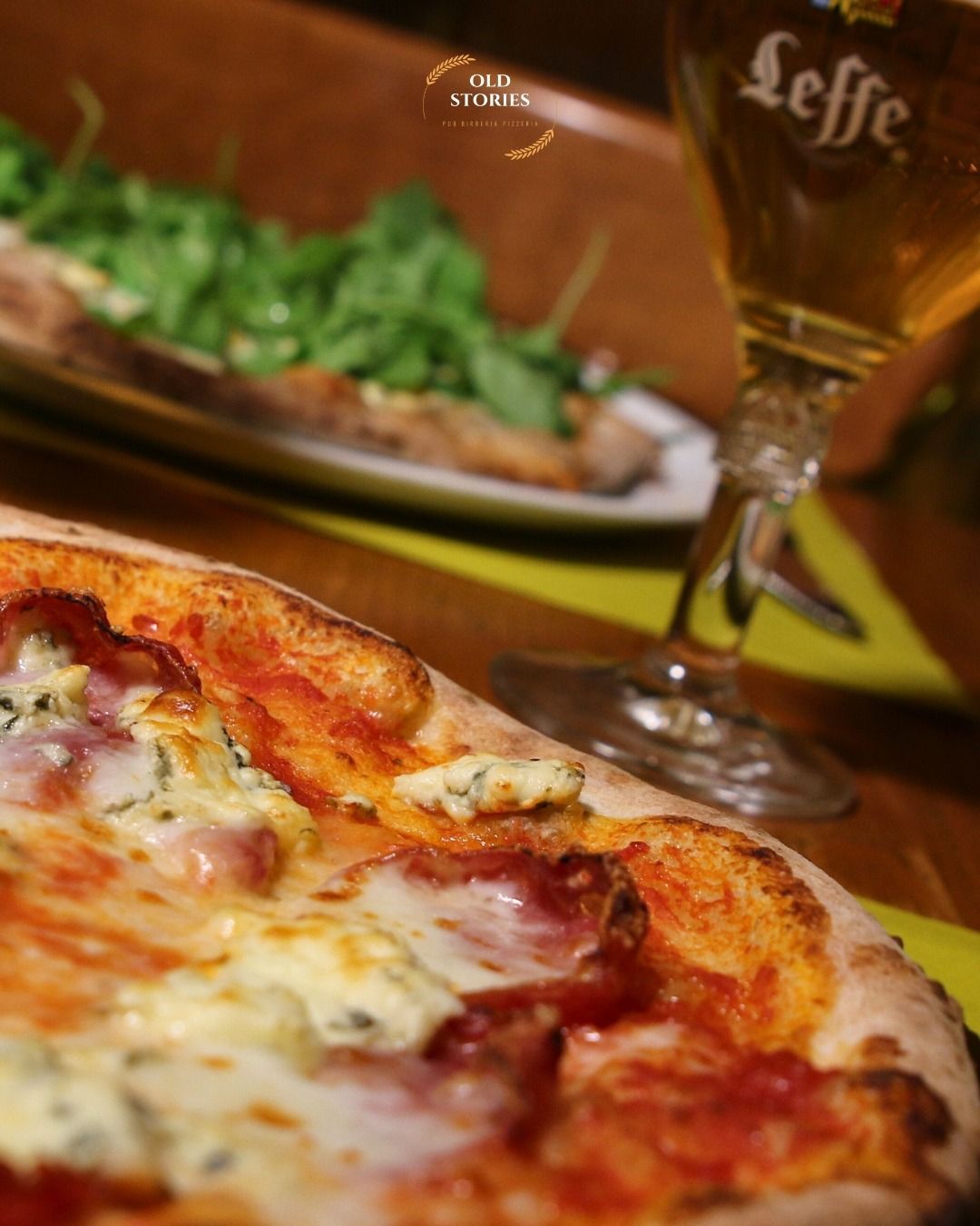 Primo piano di una pizza con gorgonzola, salame e una birra, con un piatto di rucola sullo sfondo.