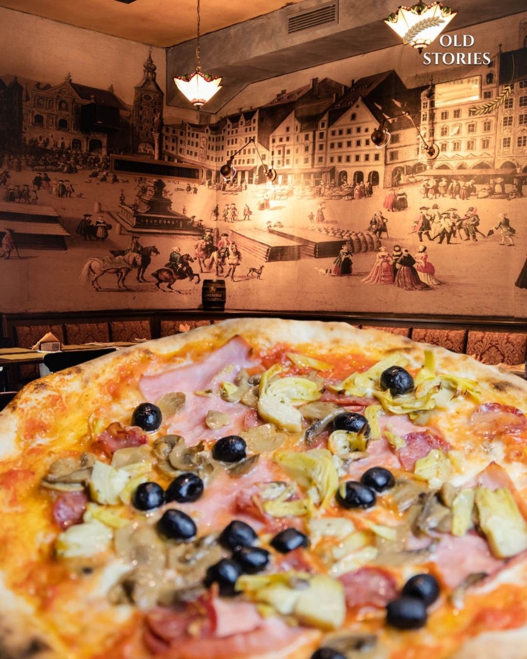 Primo piano di una pizza con prosciutto, carciofi, funghi e olive. In un ristorante con un murale sullo sfondo.