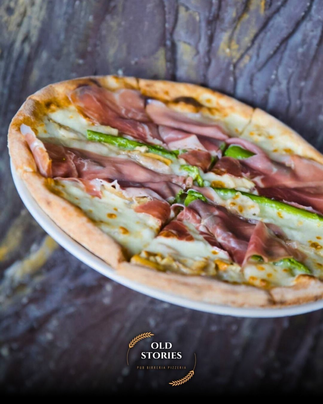 Pizza con prosciutto, asparagi e formaggio su un piatto bianco su una superficie marrone e ruvida.