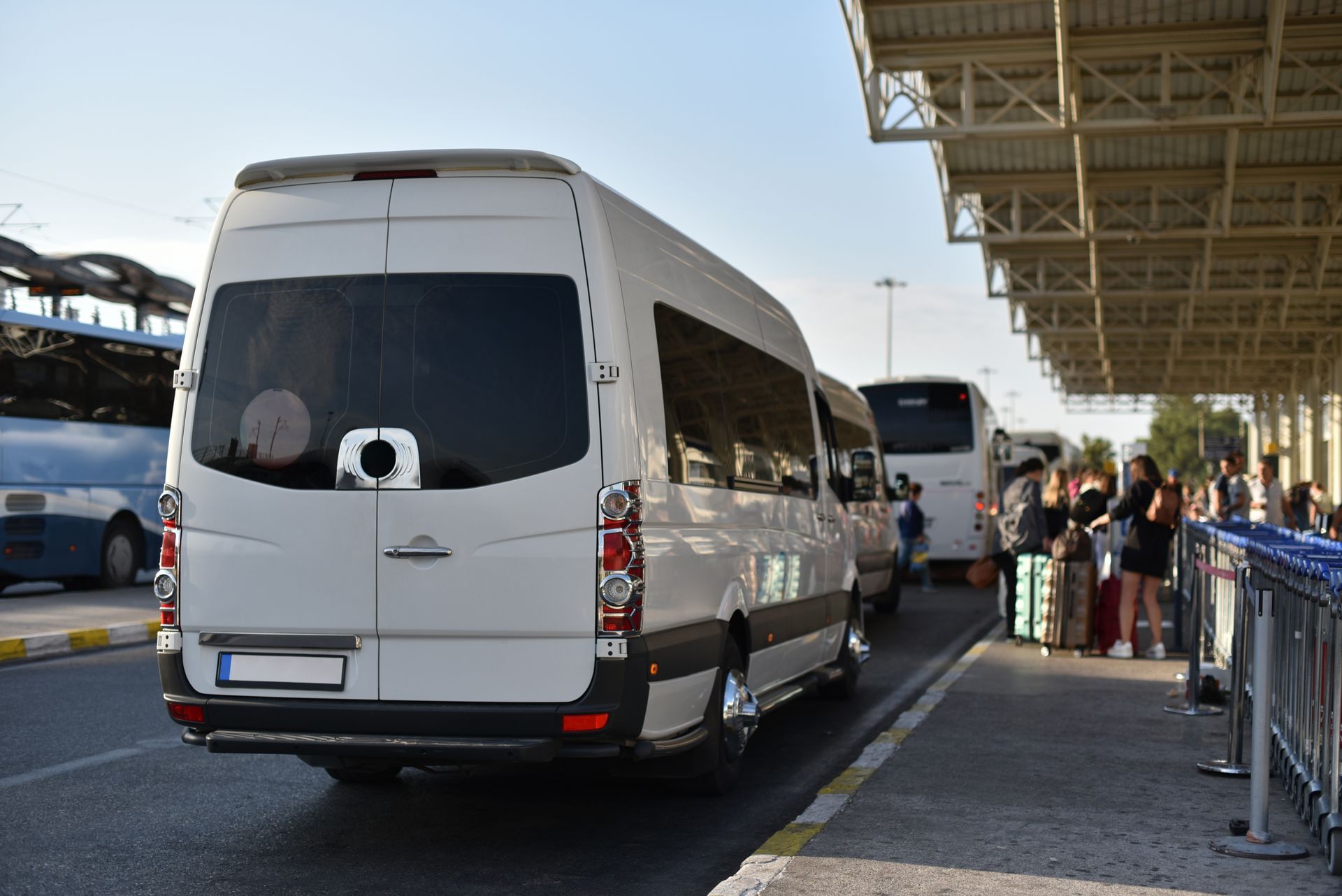 White passenger van at a bus stop, next to people with luggage; sunny outdoor setting.