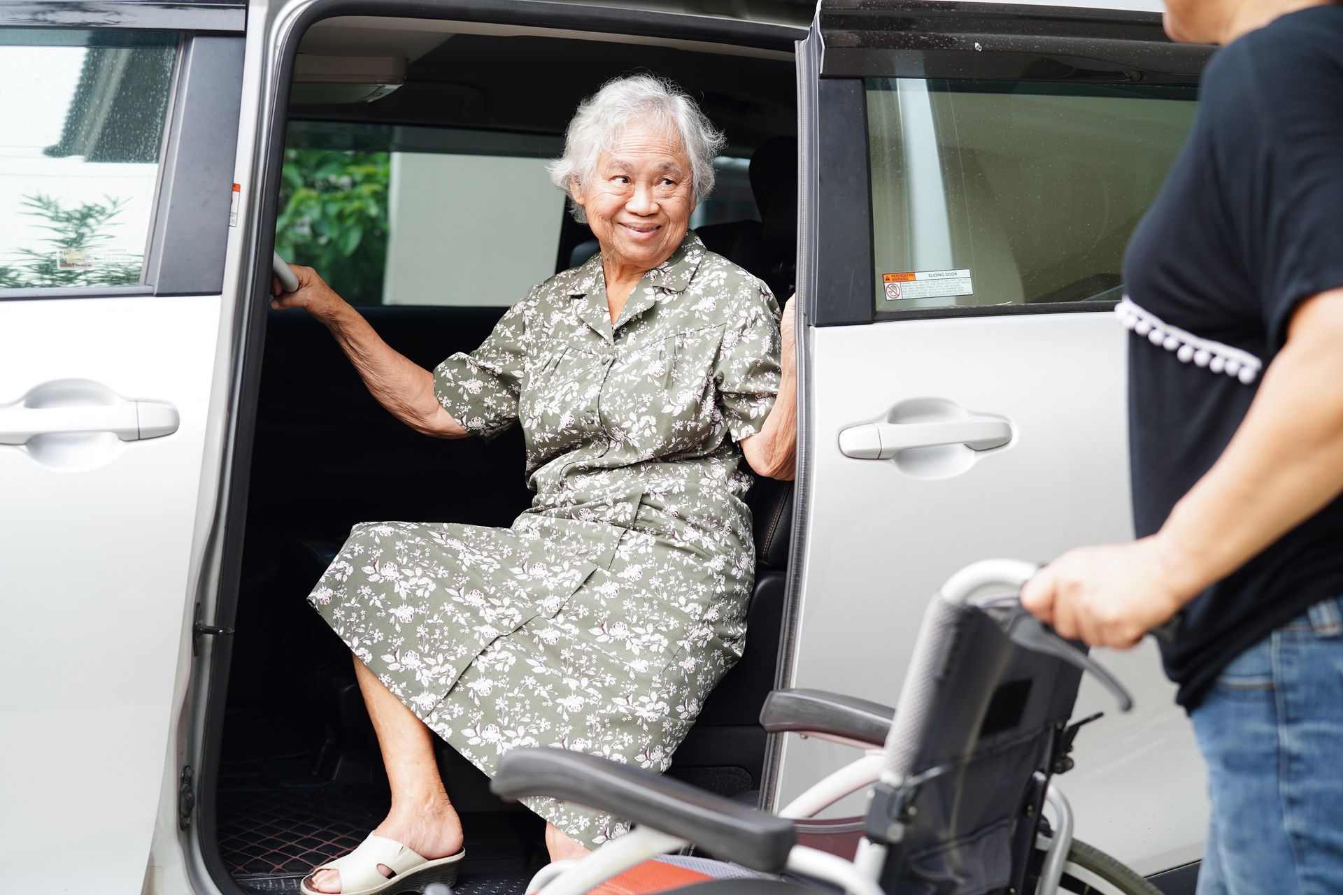 Elderly person exiting a car with assistance, wheelchair nearby.
