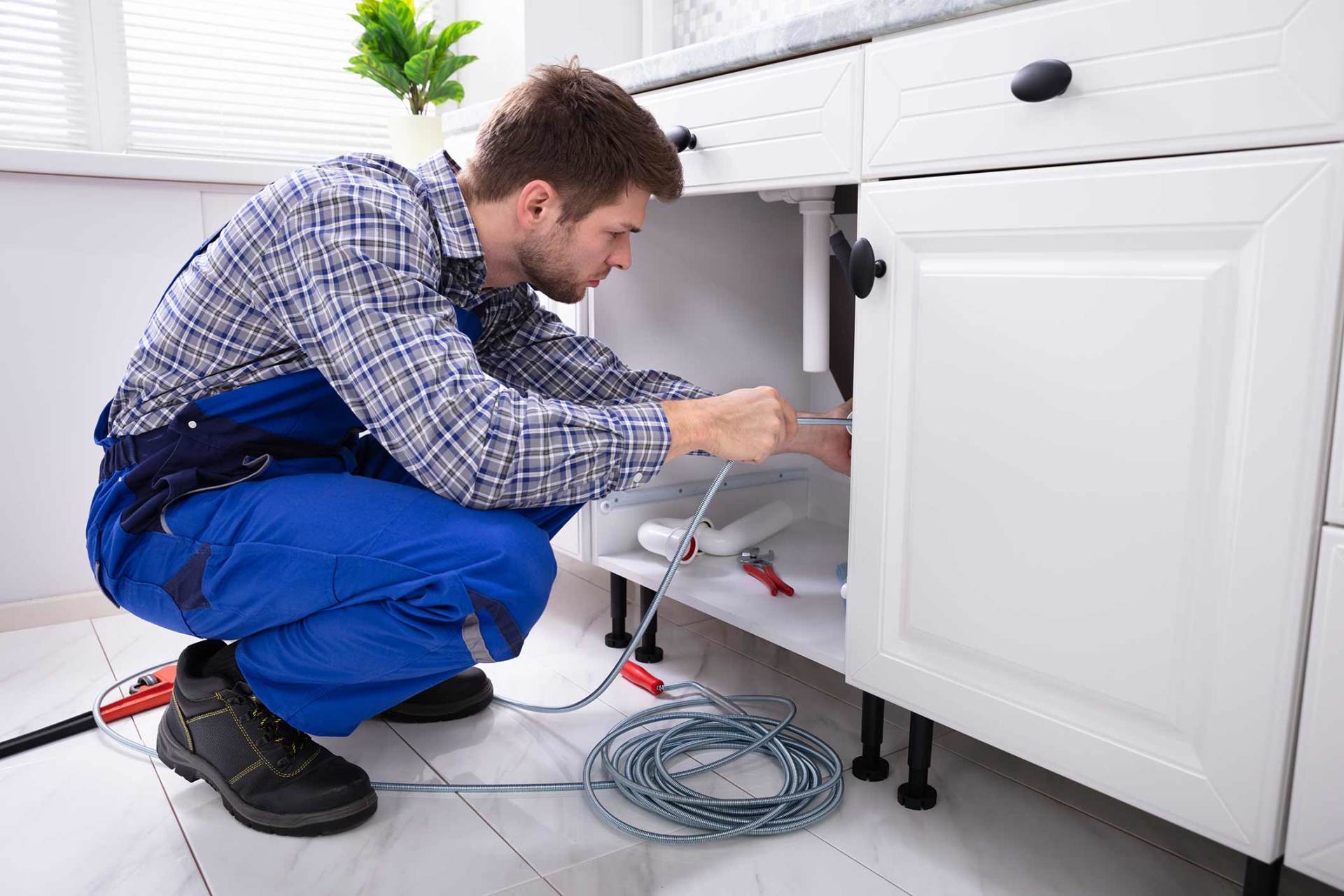 A plumber in blue overalls works under a white kitchen cabinet.