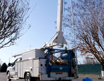 Utility truck with extended boom, blue sky, trees, white and blue truck.
