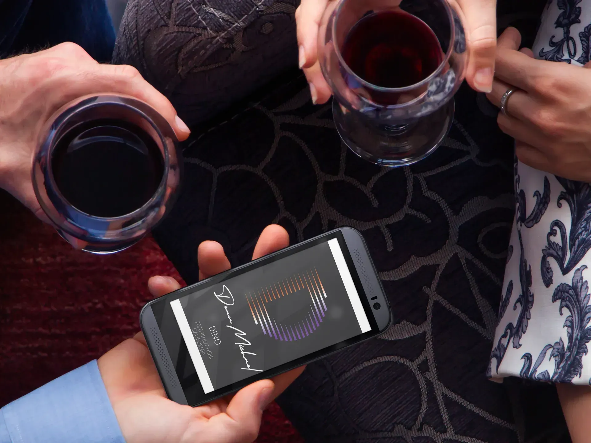 Hands holding wine glasses around a smartphone displaying a music app on a patterned couch