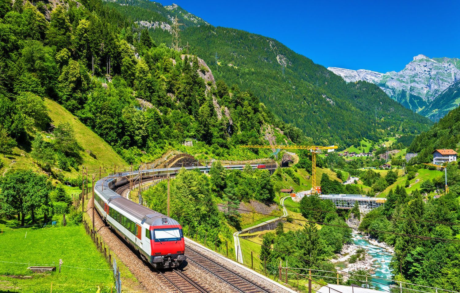 A red and white train travels along tracks winding through a vibrant, lush green mountain valley with a stream nearby.
