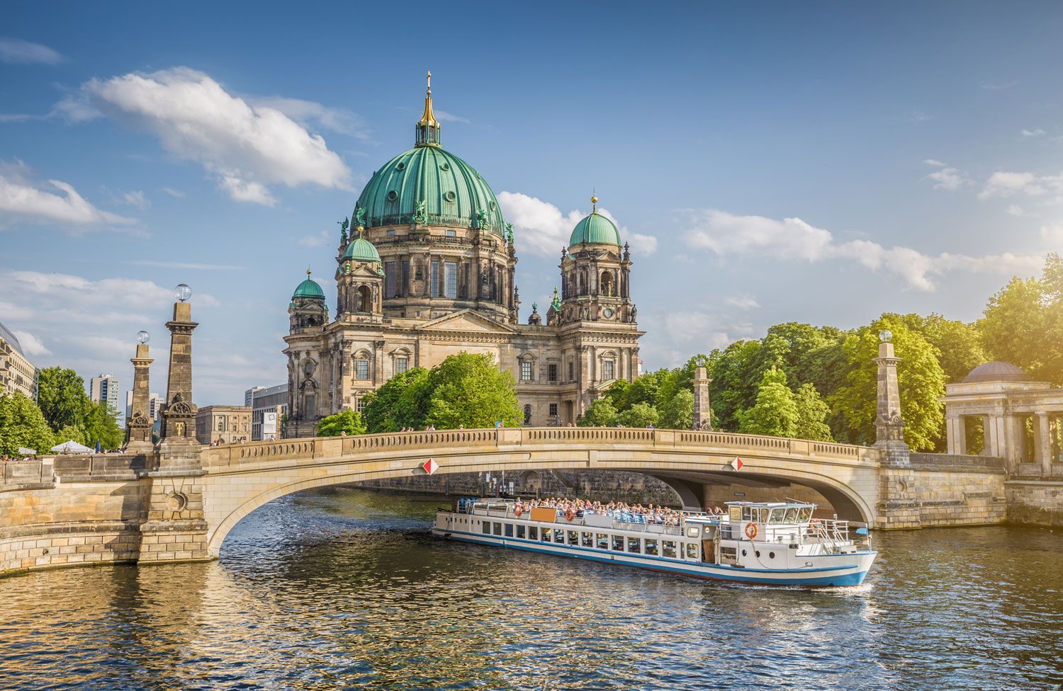 A tour boat passes under a stone bridge in front of the Berlin Cathedral with its green dome, on a sunny day.