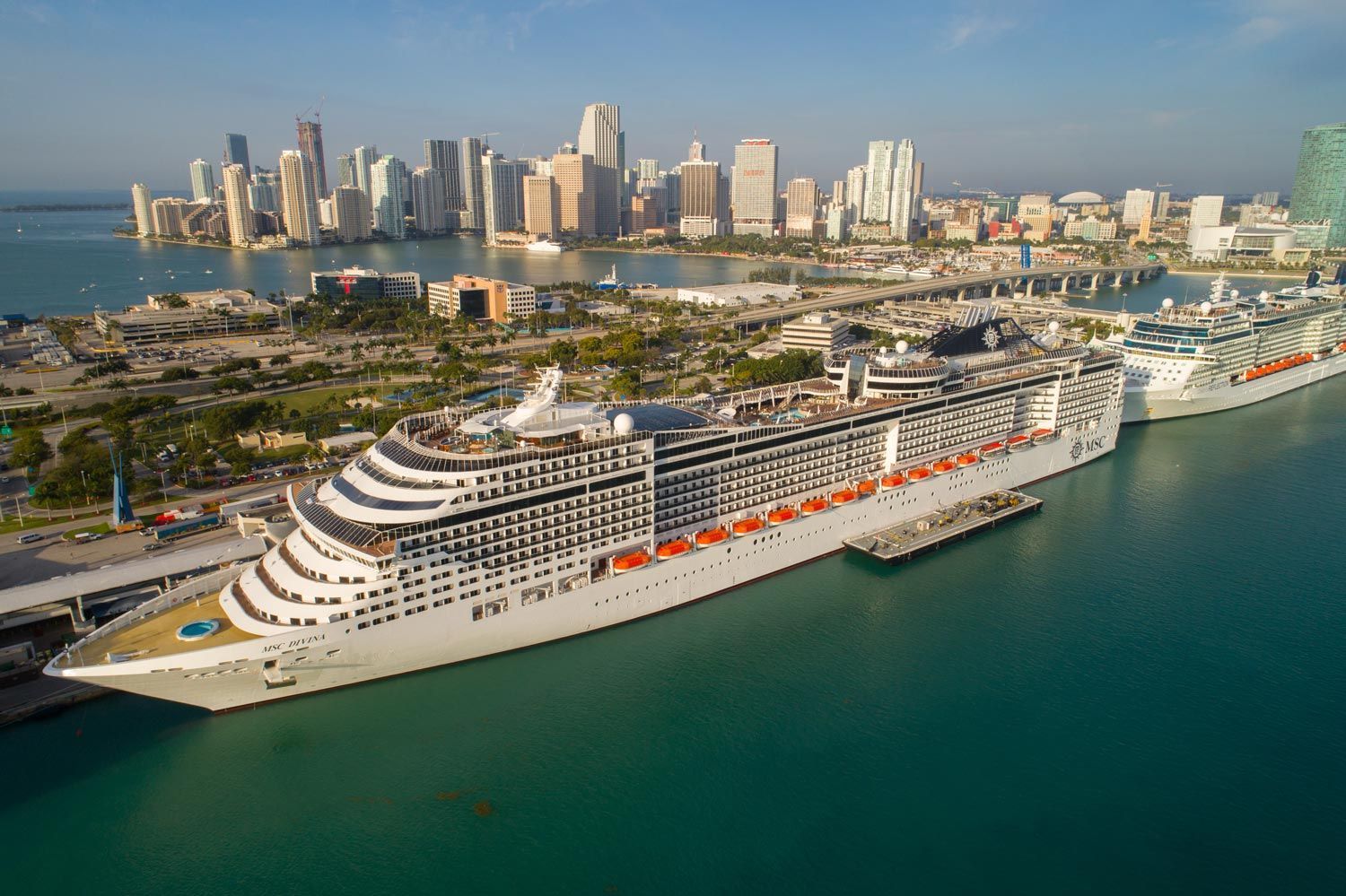A large MSC cruise ship docked at a port with the Miami skyline in the background under a clear blue sky.