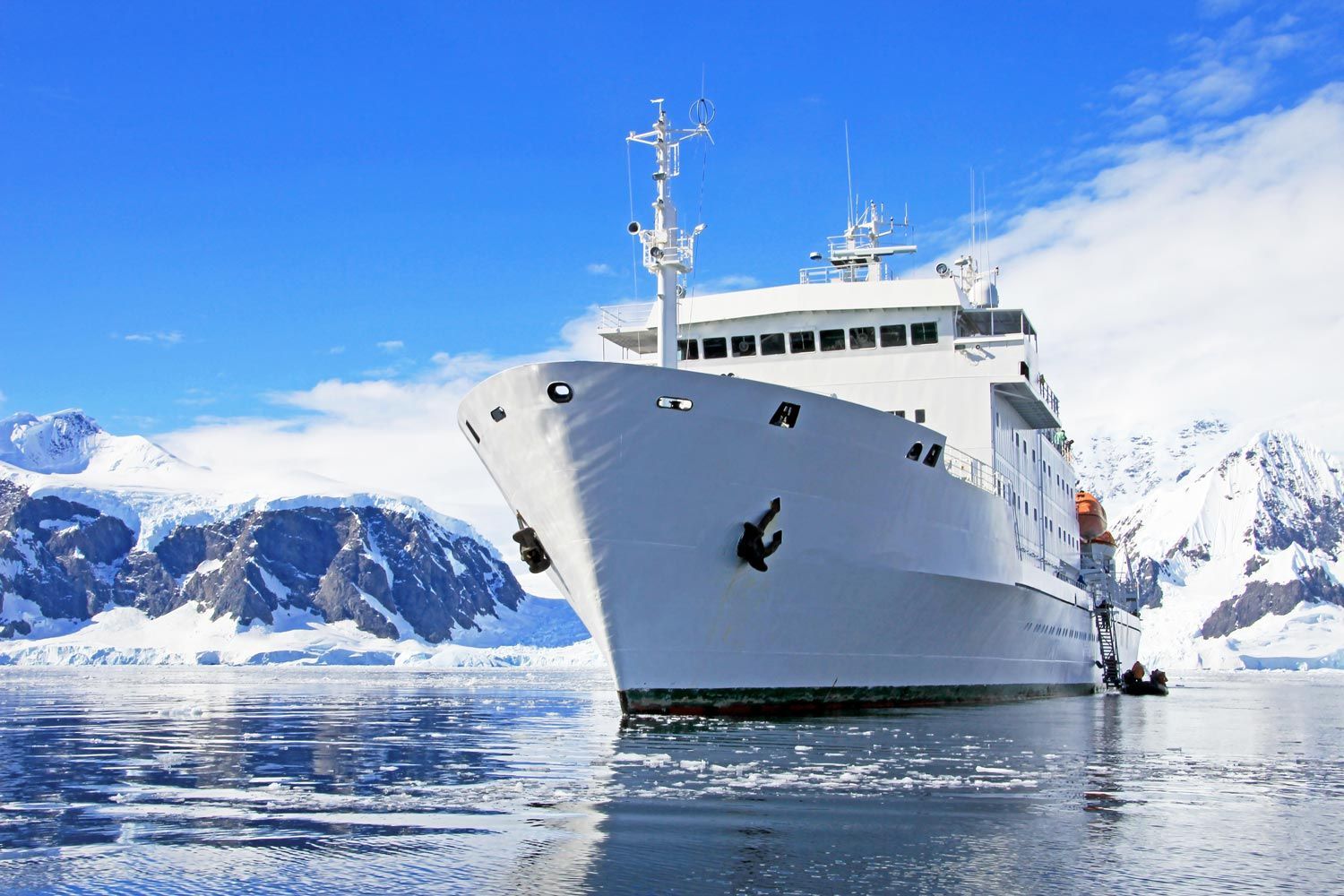 A white expedition ship floats in calm icy waters against a backdrop of snow-covered mountains under a bright blue sky. A white expedition ship floats in calm icy waters against a backdrop of snow-covered mountains under a bright blue sky.