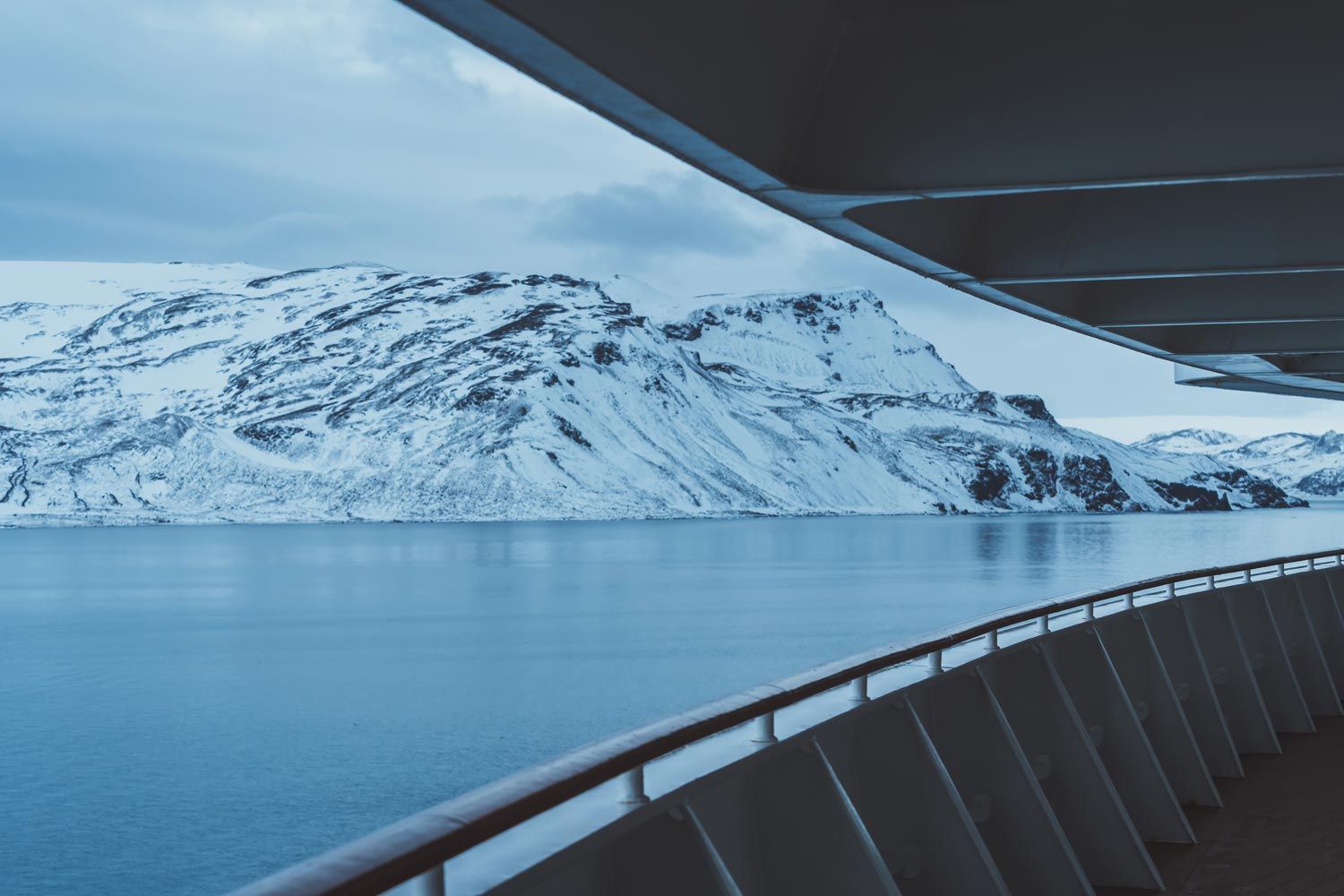 View of snow-covered mountains across a calm, blue body of water, seen from a cruise ship deck.