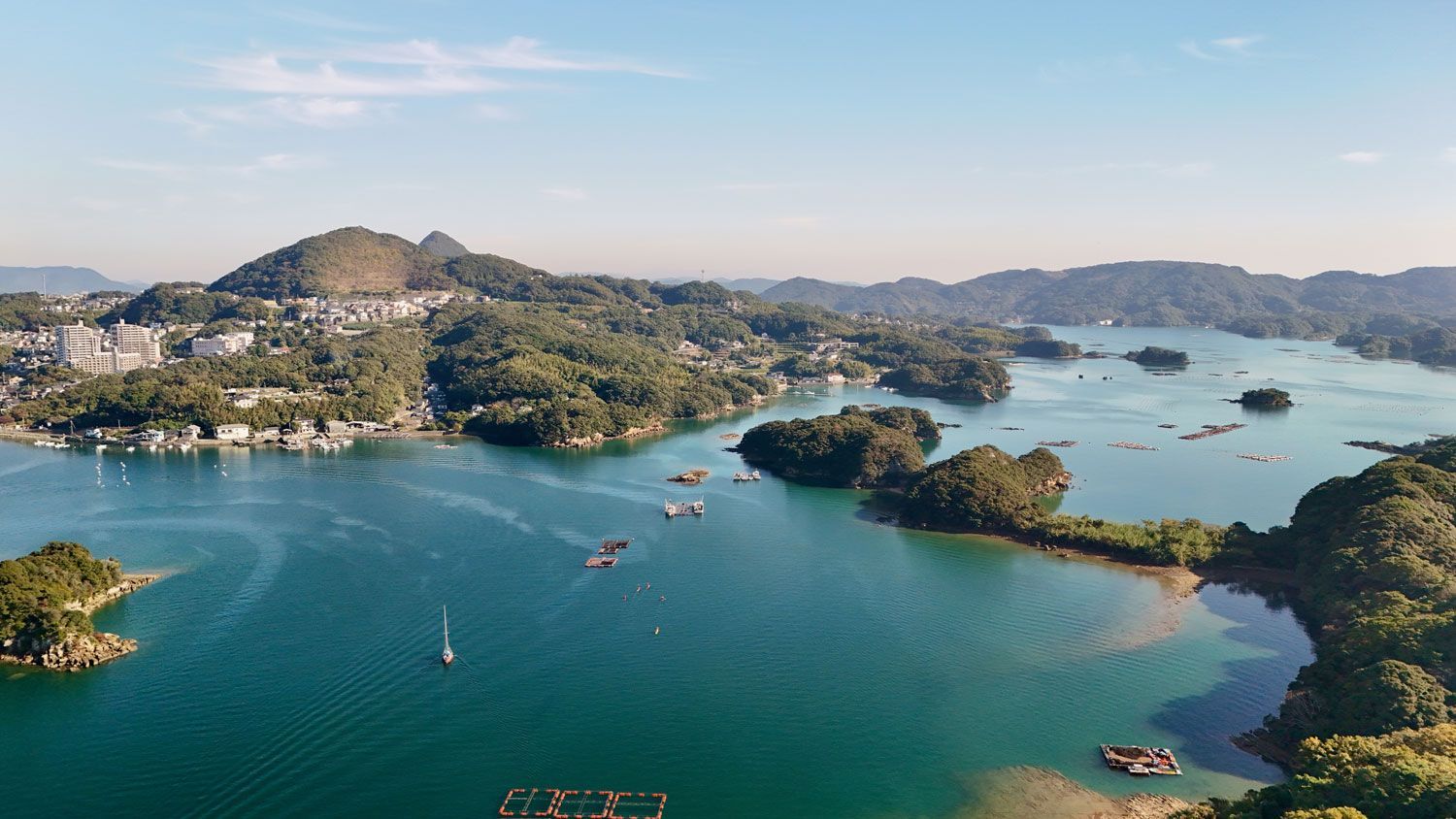 Aerial view of a calm, blue bay dotted with forested islands, small boats, and aquaculture rafts under a clear sky.