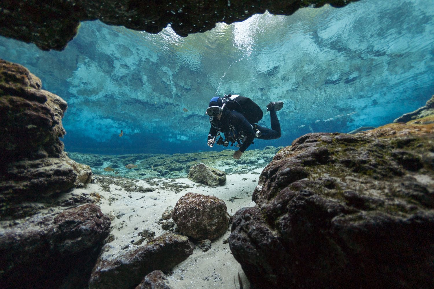 A scuba diver explores a clear, sunlit underwater cave with sandy floors and rugged rock formations.
