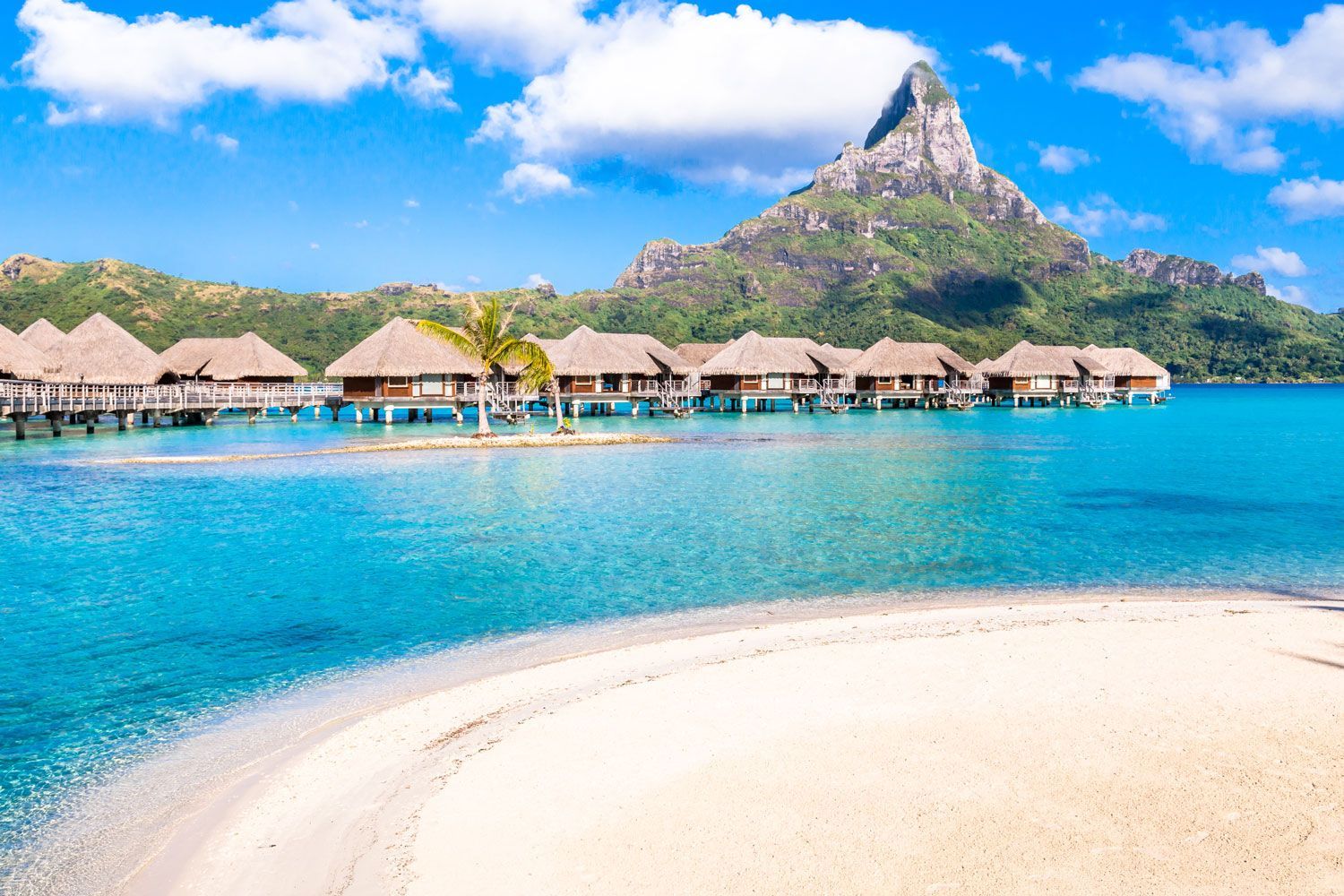 Overwater bungalows in a turquoise lagoon with a lush green mountain rising in the background under a blue sky.