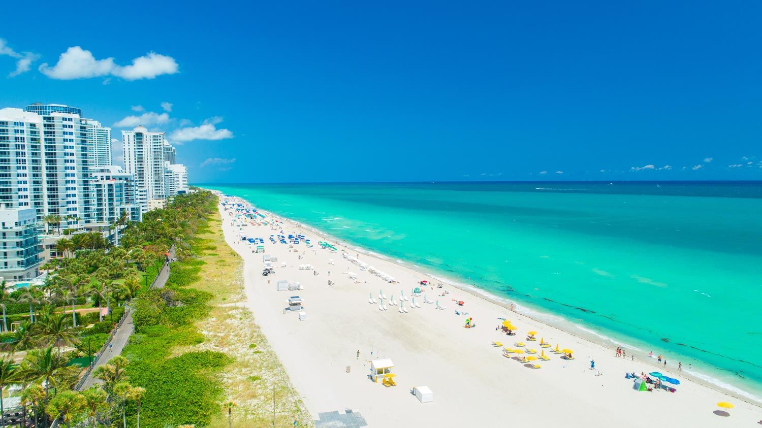 A sunny, wide beach with turquoise water, white sand, and beachfront high-rise buildings on a clear day. A sunny, wide beach with turquoise water, white sand, and beachfront high-rise buildings on a clear day.