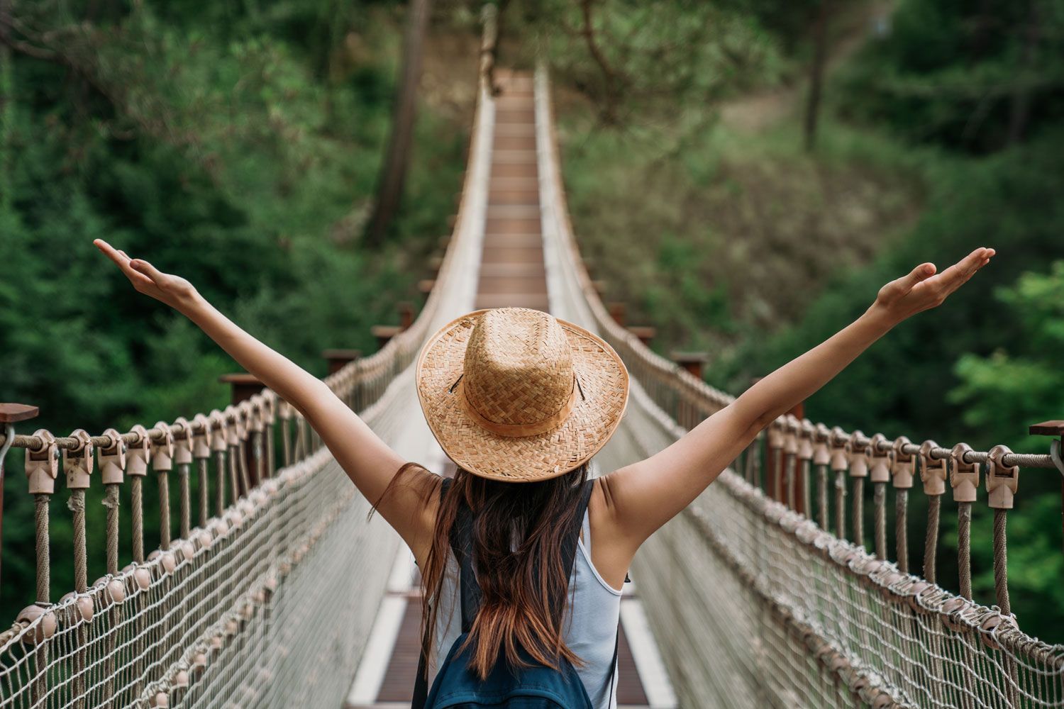 A person wearing a straw hat and backpack stands on a suspension bridge with arms outstretched, facing a lush forest.