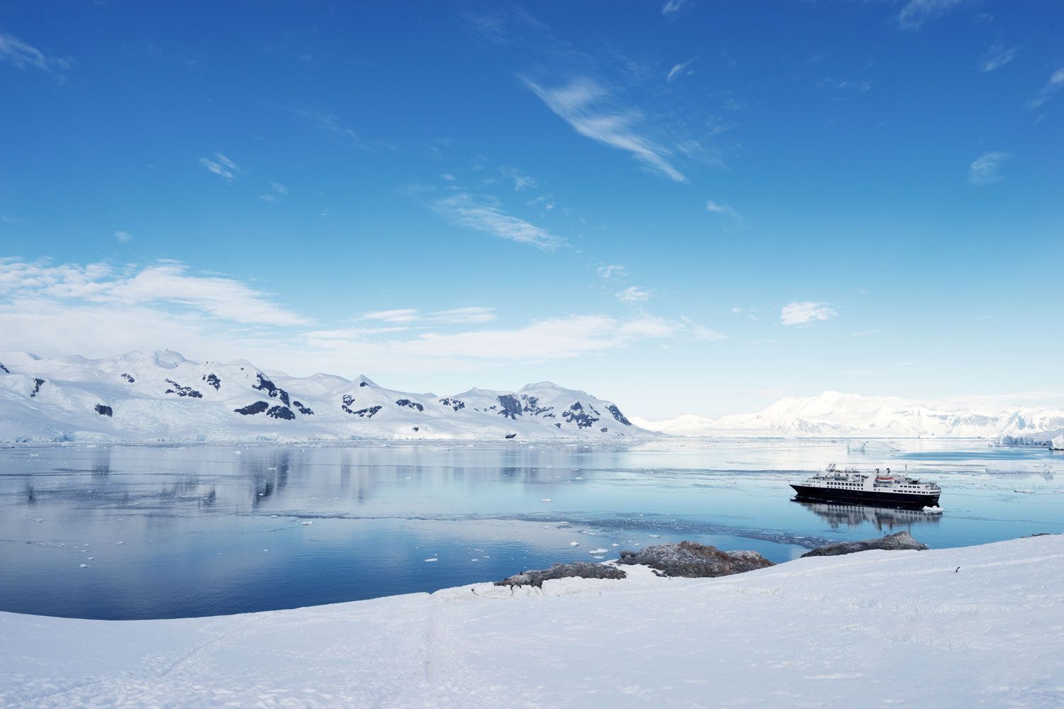 A cruise ship floats in a calm, icy bay surrounded by snowy mountains under a bright blue sky.