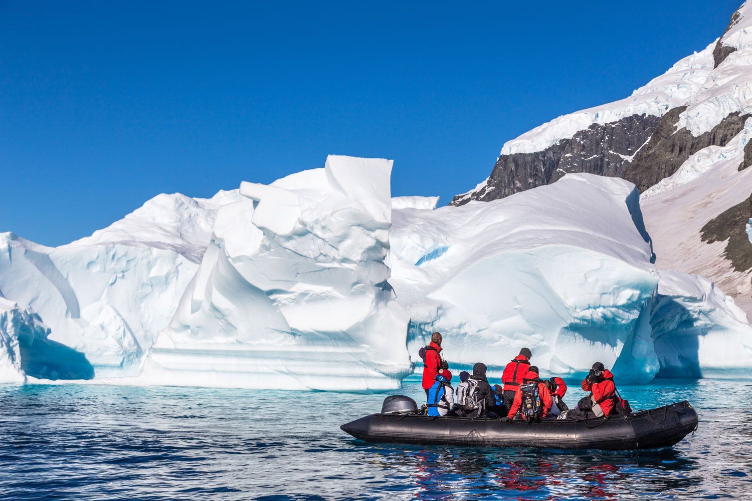 A group of people in red parkas riding a black inflatable boat past massive icebergs in a sunny, snowy landscape.