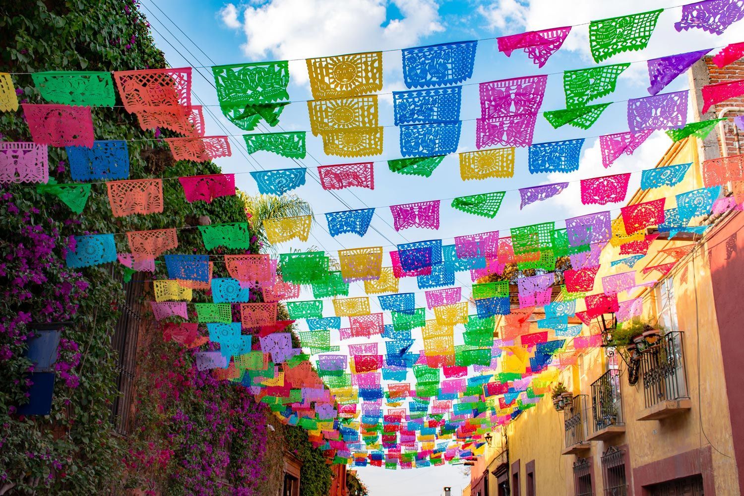 Bright, colorful Mexican paper cut-out banners, or papel picado, hang in a grid above a narrow, sunlit colonial street.